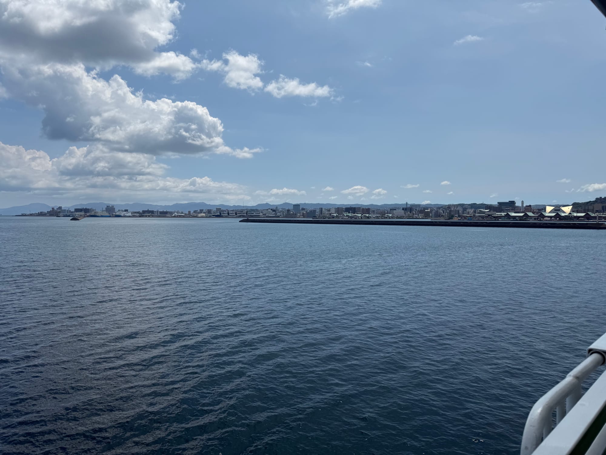 View from a ferry crossing Kagoshima Bay towards the city of Kagoshima, with calm blue water, the waterfront skyline and dramatic summer clouds overhead.