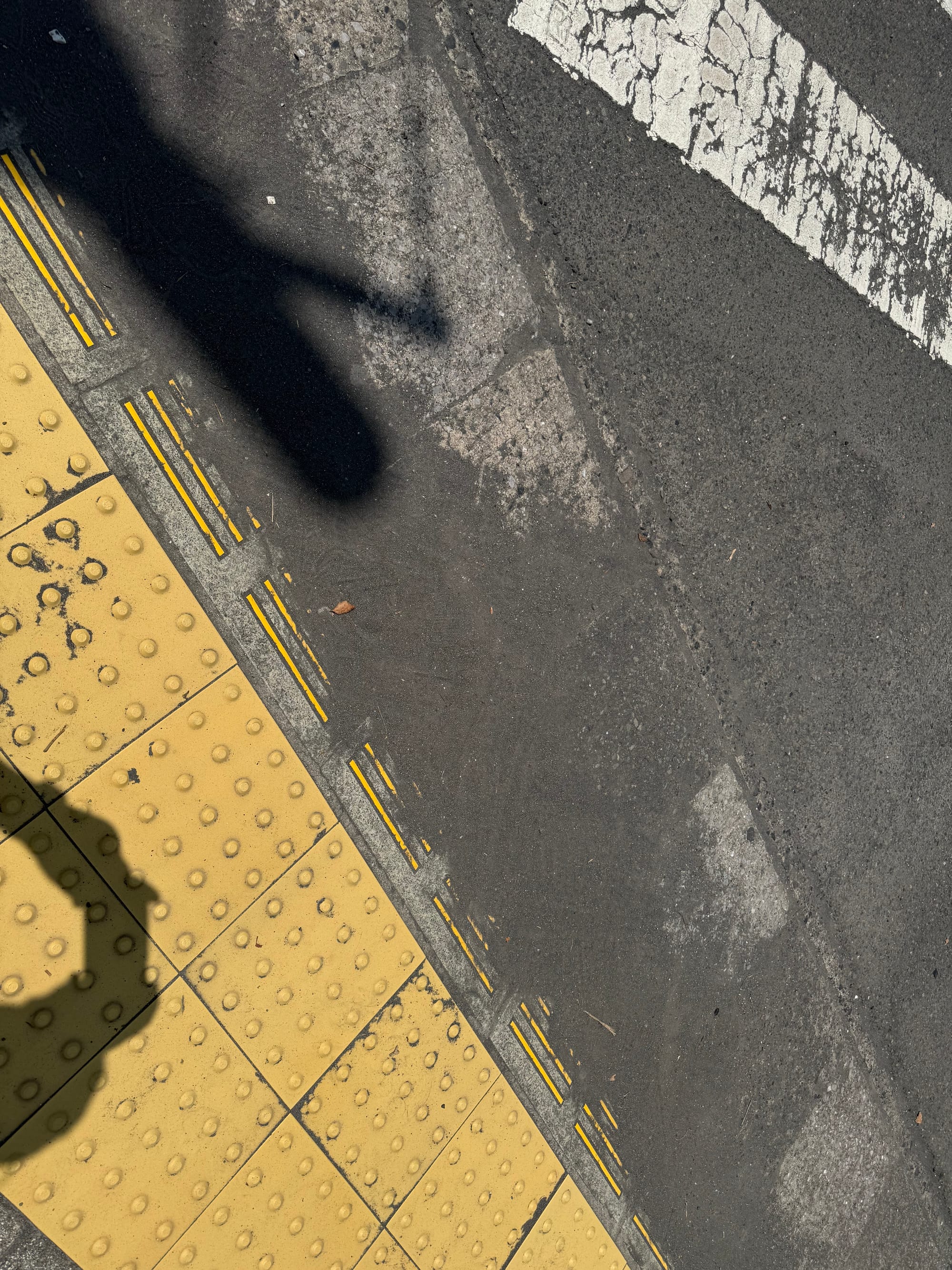 Volcanic ash accumulated along the roadside on Sakurajima, visible beside yellow tactile paving and a pedestrian crossing, showing the effects of ongoing volcanic activity.