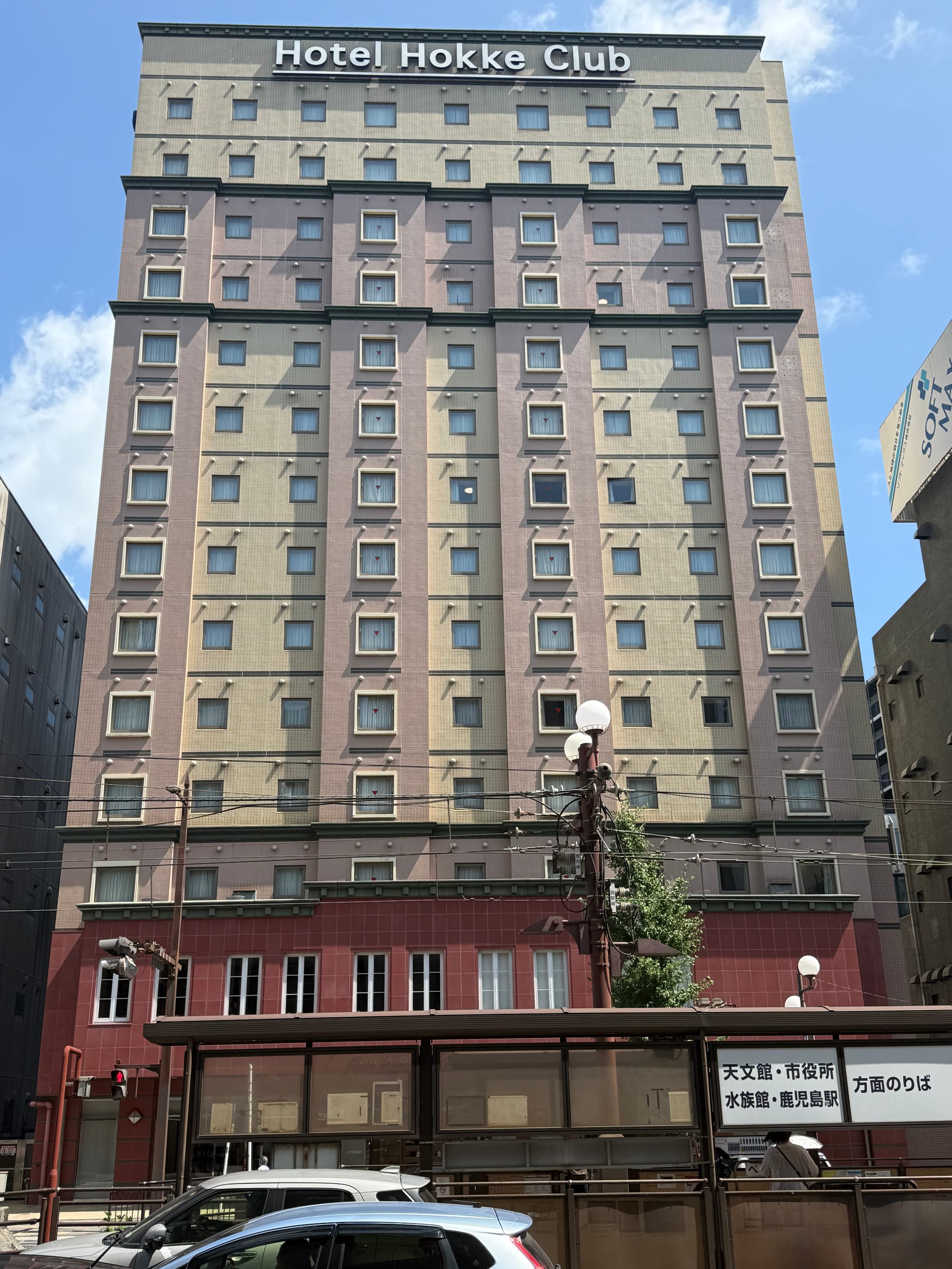 Hotel Hokke Club Kagoshima, a multi-storey hotel building in central Kagoshima, viewed from across the street beneath a bright blue summer sky.