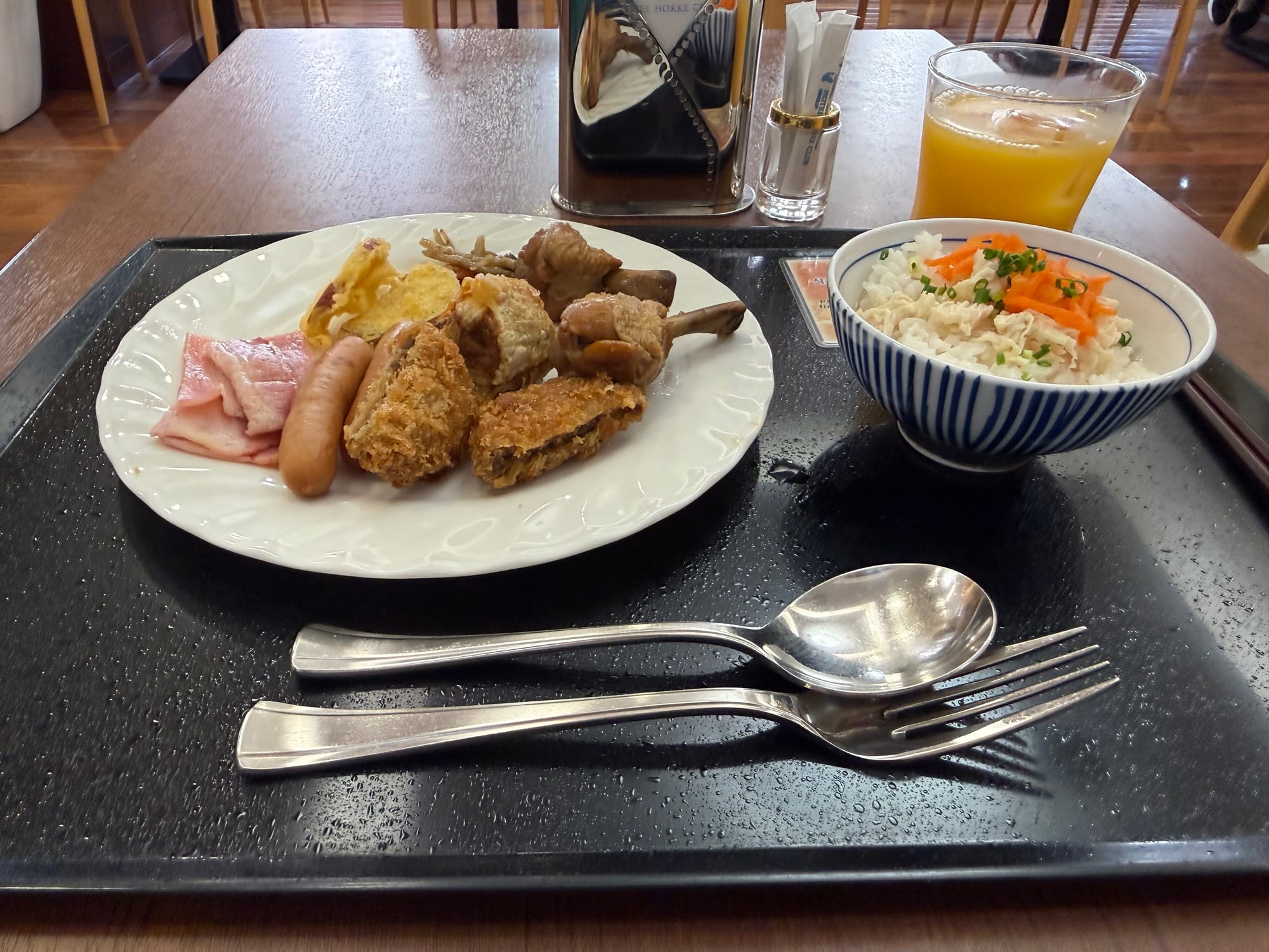 Hotel breakfast in Kagoshima featuring sausages, ham, fried items, braised pork, potato dishes, shredded chicken salad and a glass of orange juice.