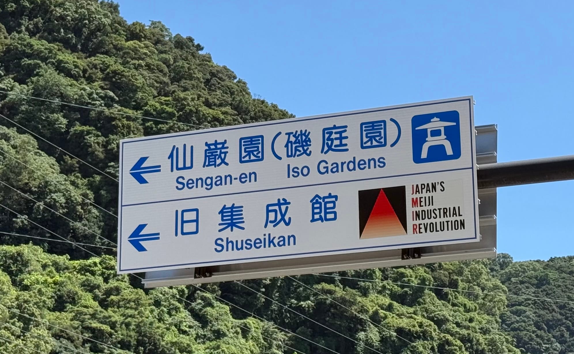 Directional sign near Sengan-en in Kagoshima showing routes to Sengan-en (Iso Gardens) and the Shuseikan museum, with wooded hills rising behind.