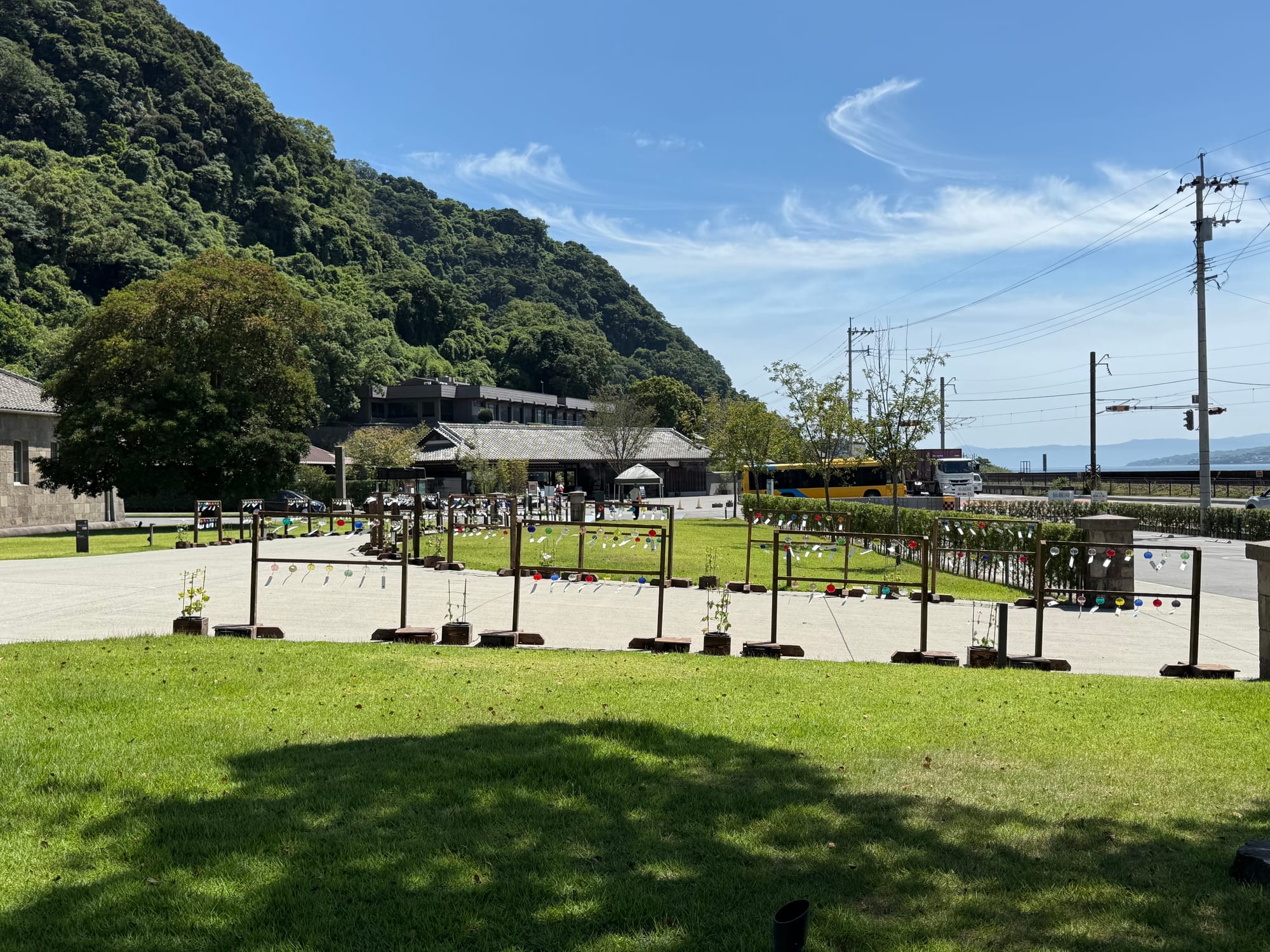 Lawn and visitor area at Sengan-en in Kagoshima, with colourful glass decorations, historic buildings, wooded hills and Kagoshima Bay visible beyond.