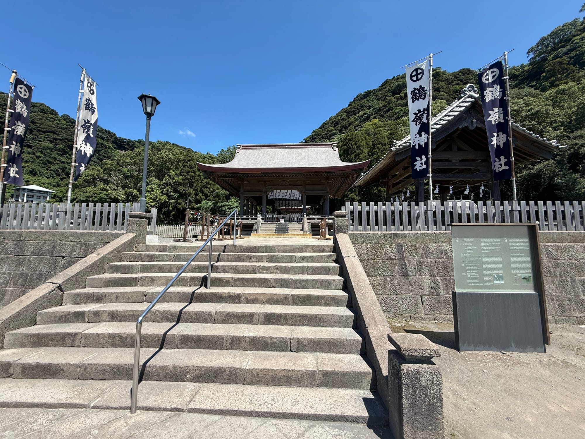 Stone steps lead up to Tsurugane Shrine at Sengan-en, with traditional shrine buildings, banners and forested hills forming a dramatic backdrop beneath a clear blue sky.