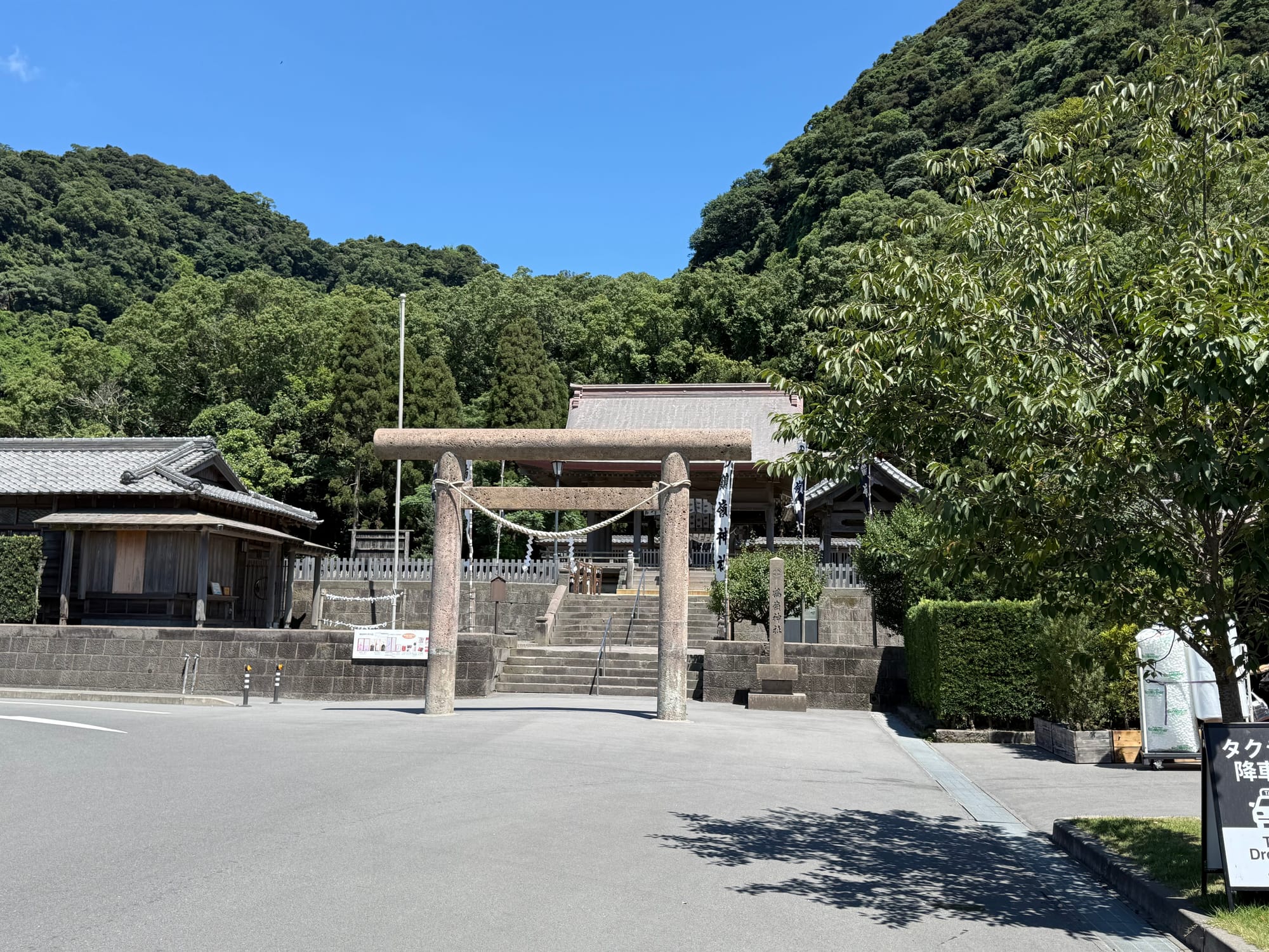 Stone torii gate at Sengan-en in Kagoshima, standing before a Shinto shrine and stone steps, with forest-covered hills rising behind beneath a clear blue sky.
