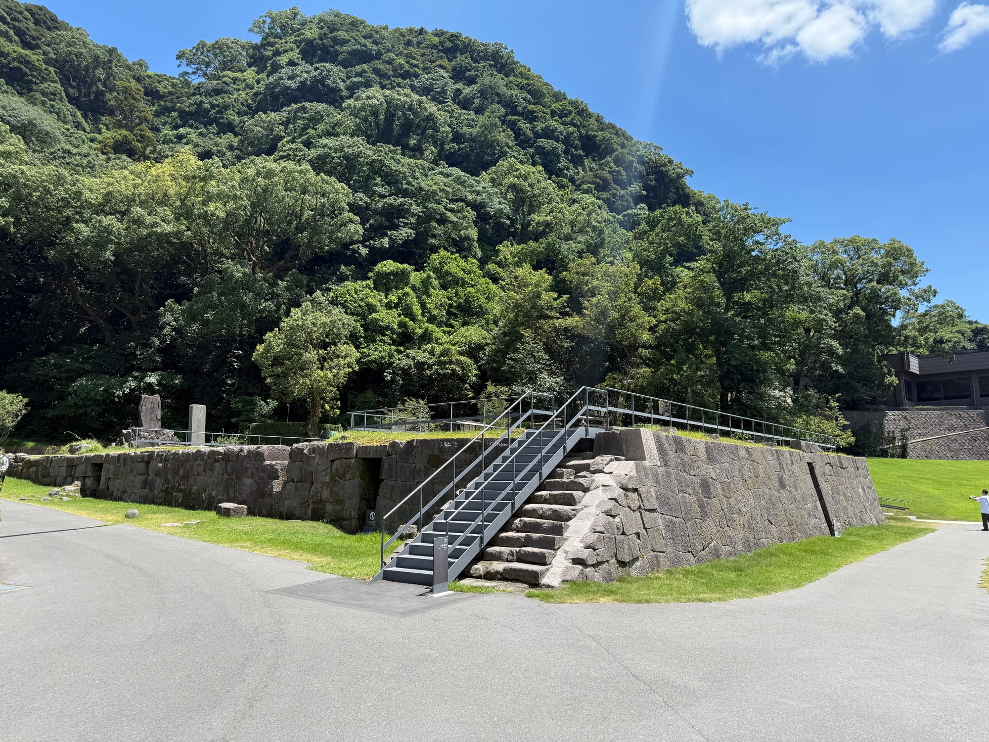 Site of the reverberatory furnace at Sengan-en, featuring preserved stone foundations, a viewing platform and forested hills rising behind beneath a bright blue sky.