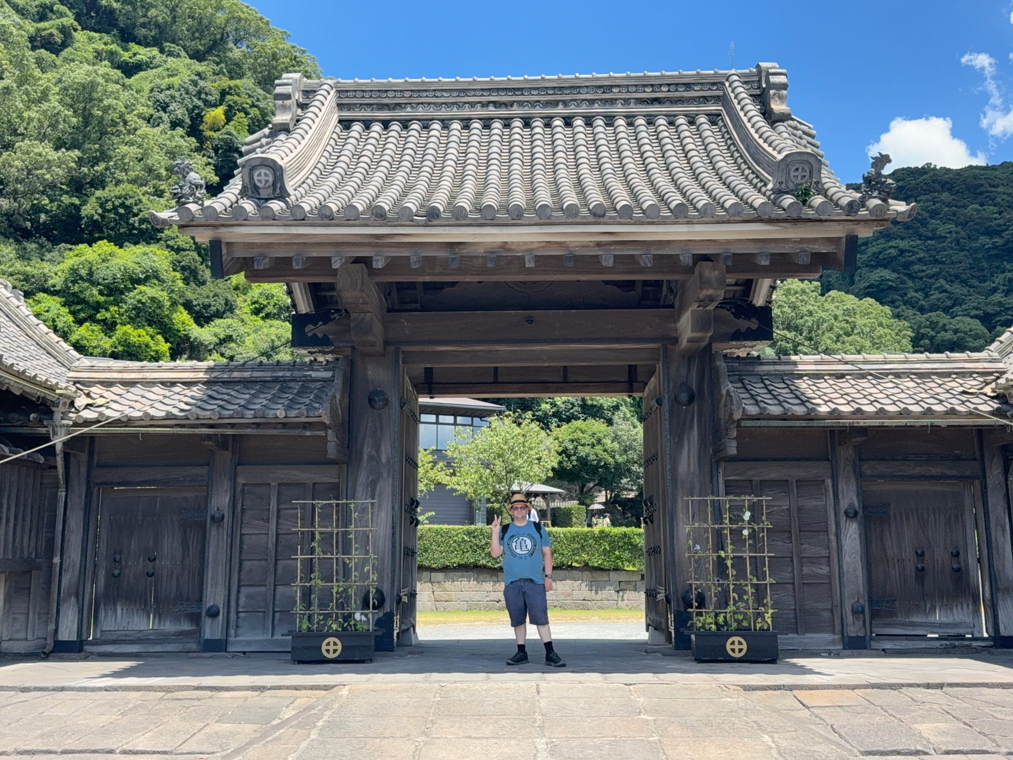 Visitor stands beneath the traditional wooden gate at Sengan-en in Kagoshima, making a peace sign, with forested hills and historic architecture in the background.