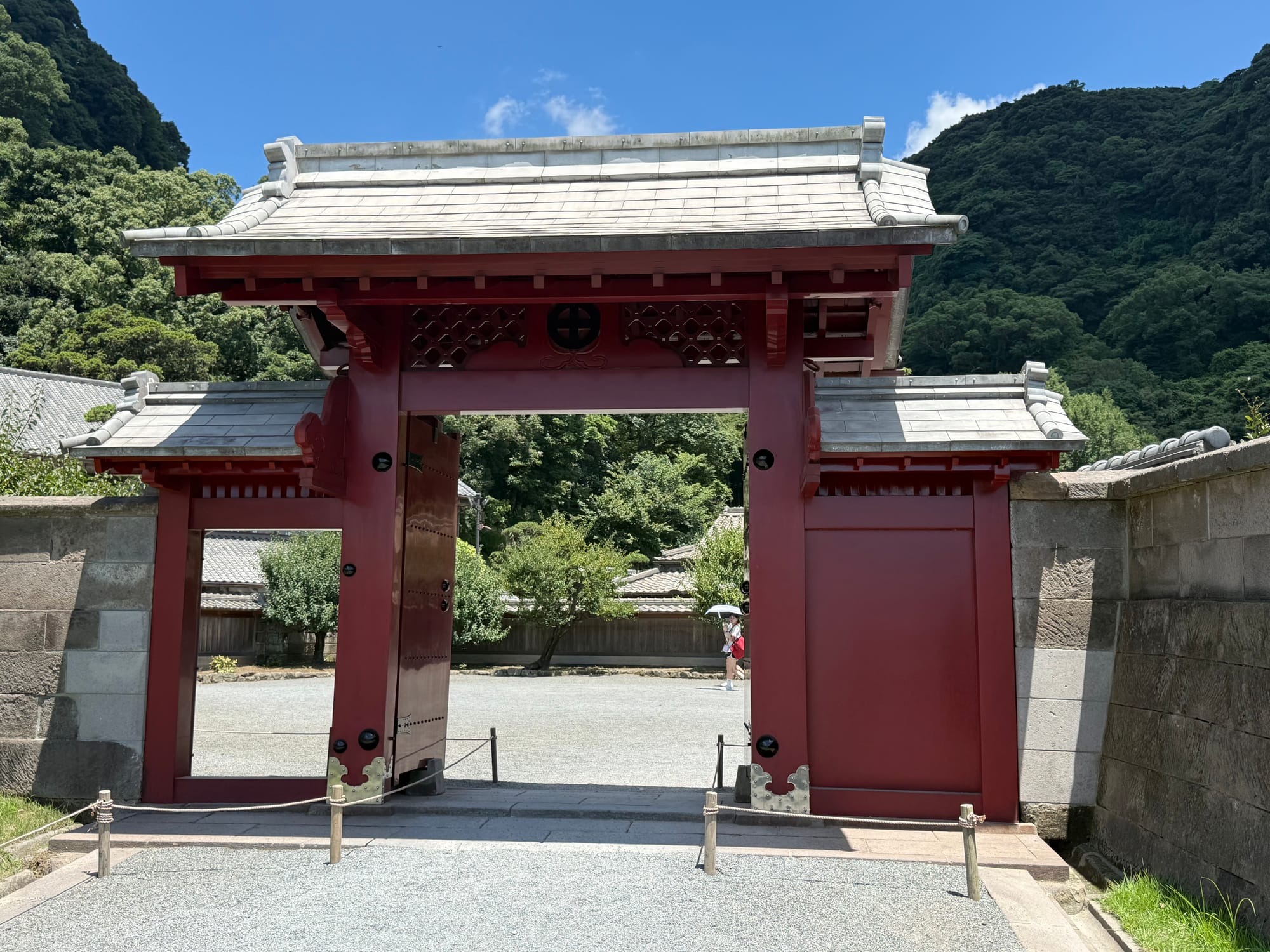 Red gate at Sengan-en in Kagoshima, featuring traditional Japanese architecture, white roof tiles and stone walls, with forested hills rising behind under a bright blue sky.