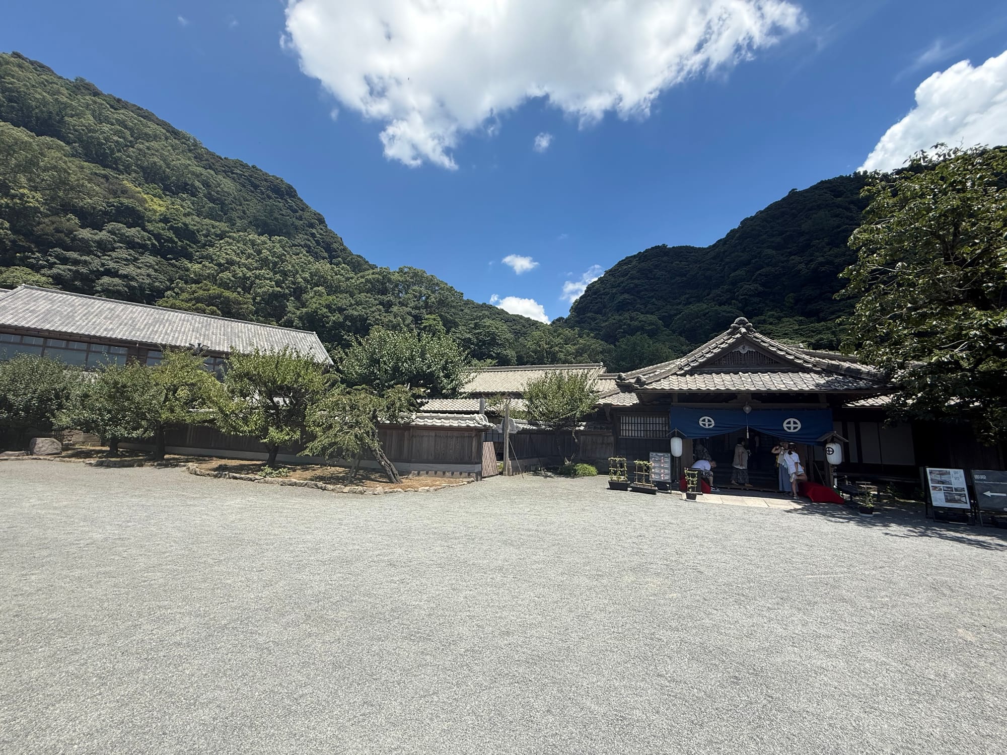Traditional buildings and a gravel courtyard at Sengan-en in Kagoshima, set against steep forested hills beneath a bright blue sky with scattered clouds.