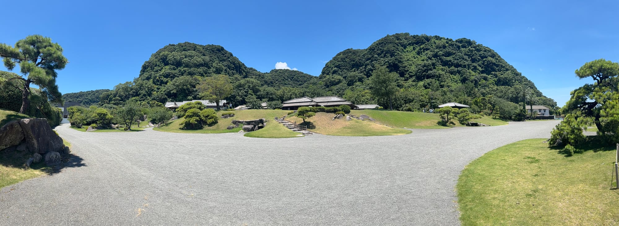 Panoramic view of the grounds of Sengan-en in Kagoshima, featuring manicured lawns, gravel pathways, traditional buildings and forested hills beneath a clear blue sky.