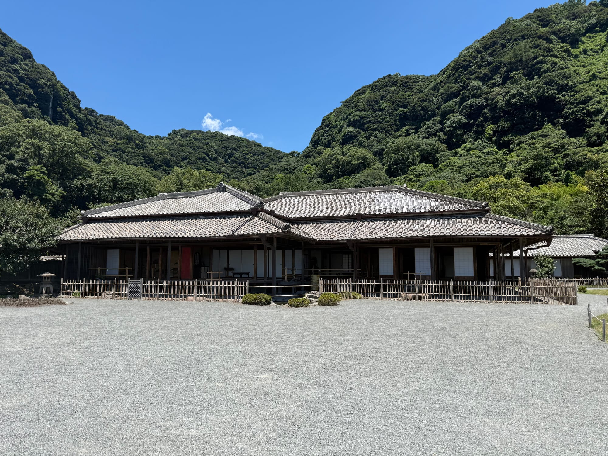 Traditional residence at Sengan-en in Kagoshima, featuring wooden architecture, tiled roofs and a wide gravel forecourt, backed by densely forested hills.