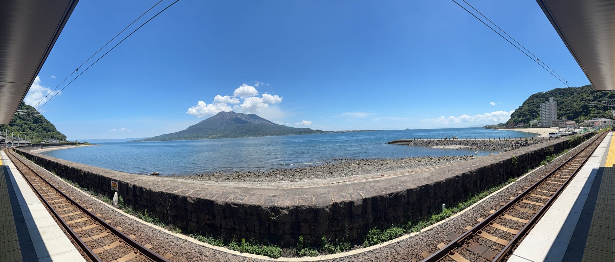 Panoramic view from Sengan-en Station, with railway tracks framing Kagoshima Bay and Sakurajima volcano rising across the water beneath a clear blue sky.