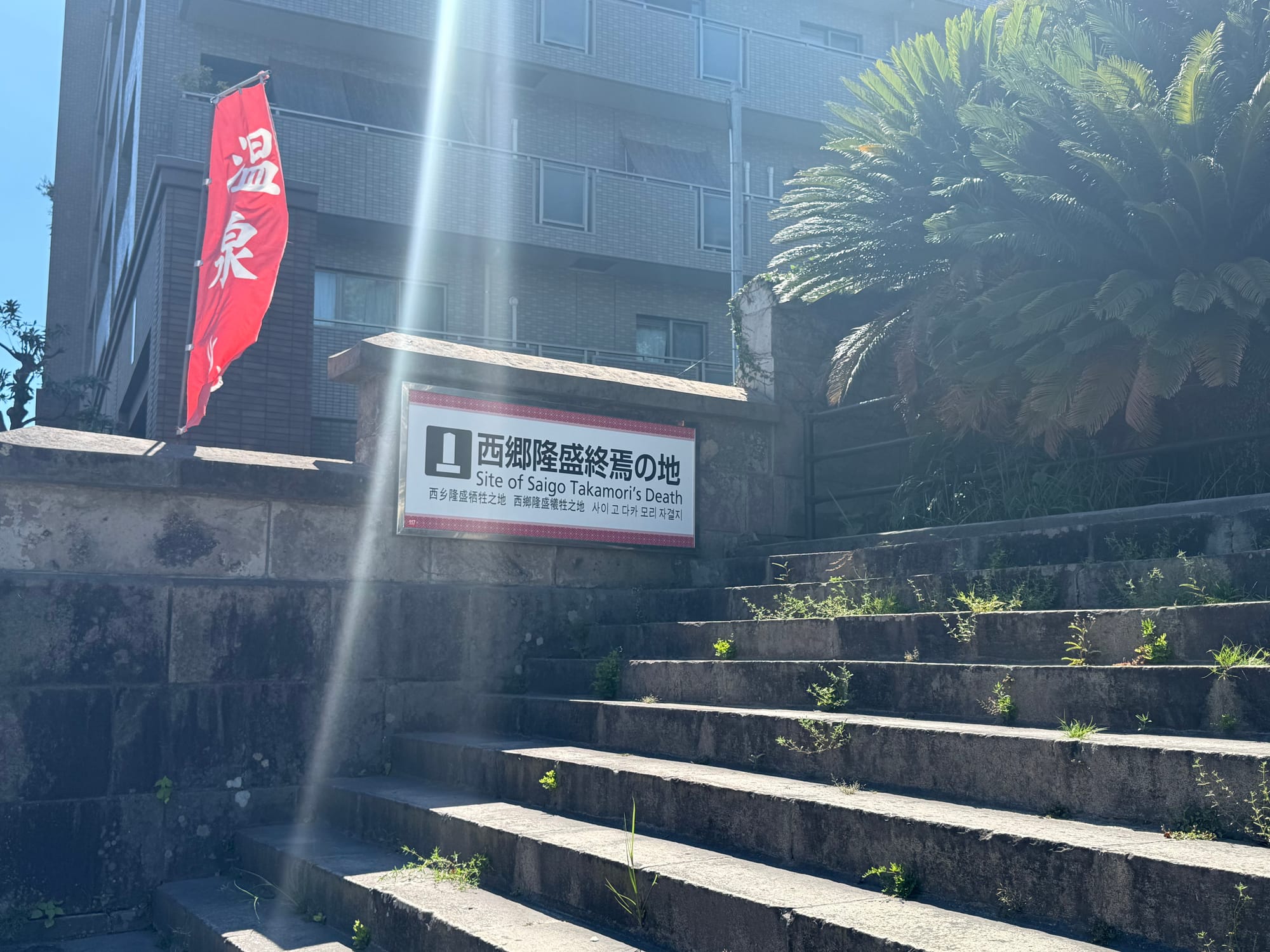 Sign marking the site of Saigō Takamori's death in Kagoshima, beside stone steps and a red banner, with sunlight streaming across the scene on a clear day.