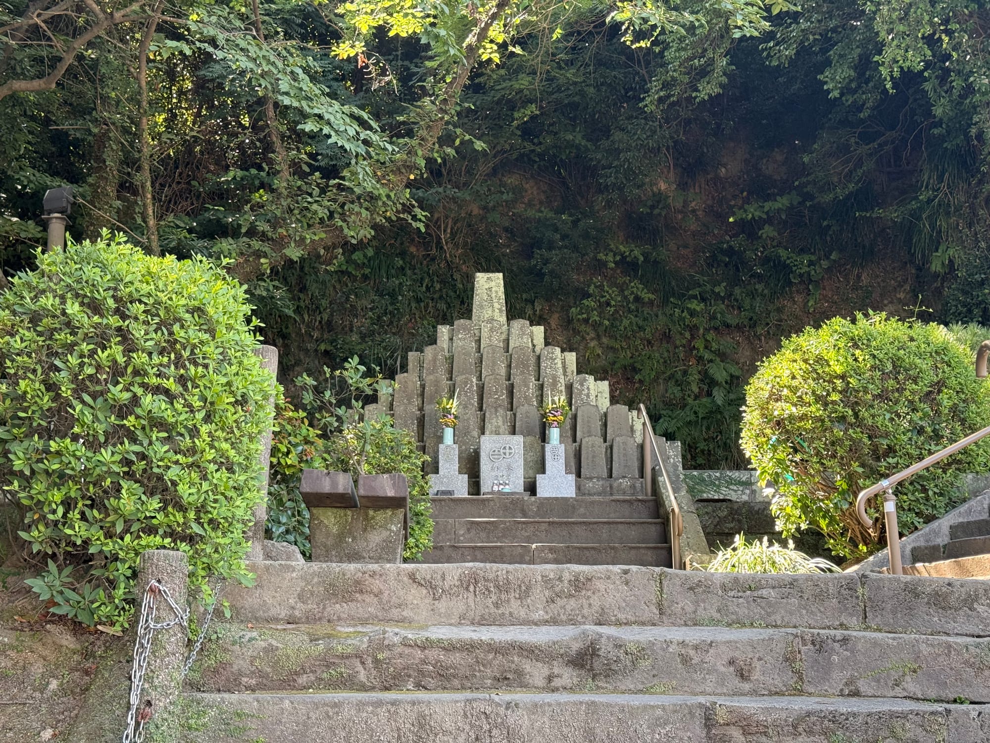 Memorial monument in Kagoshima dedicated to Saigō Takamori and his followers, standing at the top of stone steps and surrounded by trees and manicured shrubs.