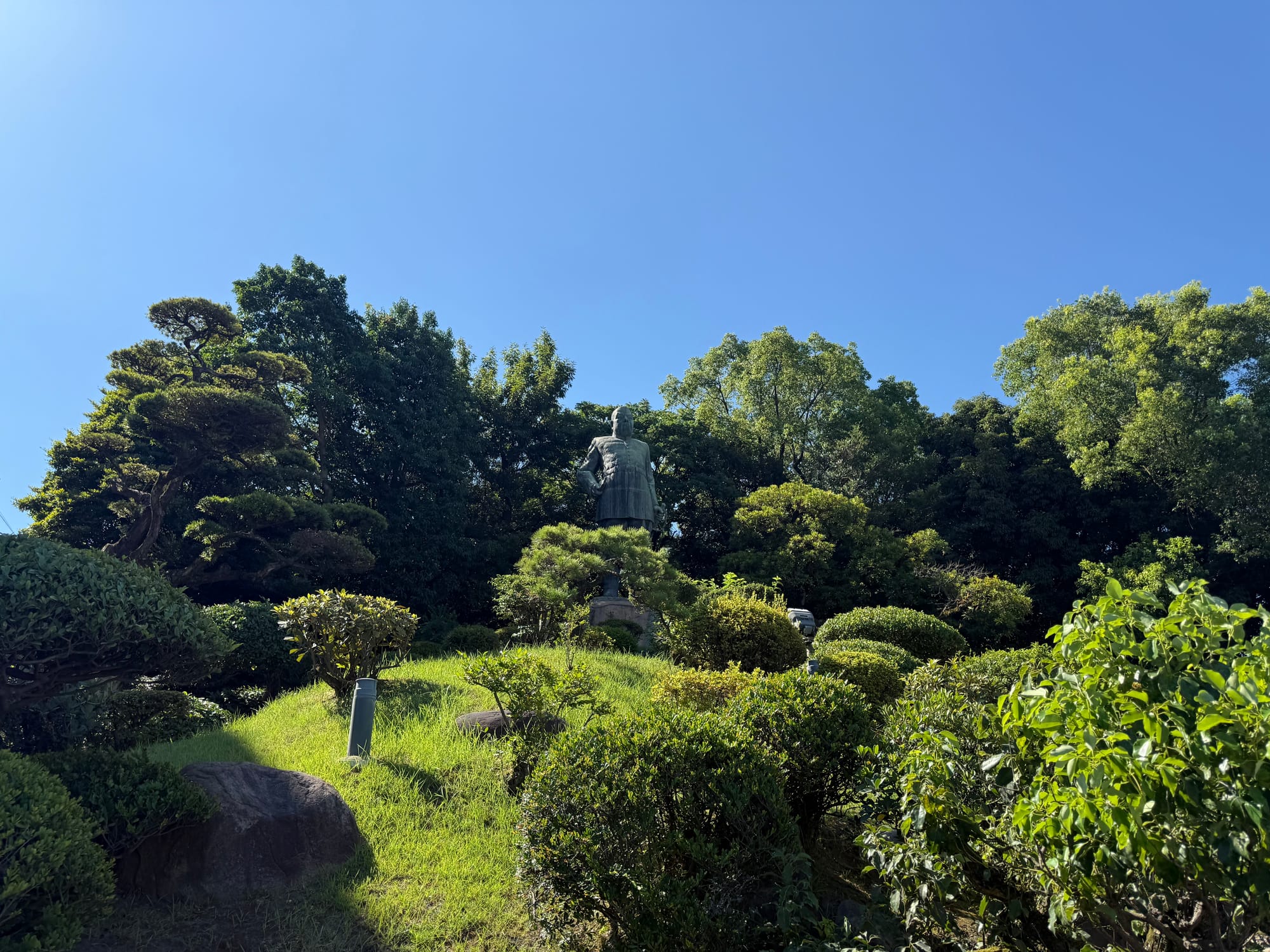 Statue of Saigō Takamori standing among landscaped gardens and trees near the Saigo Nanshu Memorial Museum in Kagoshima beneath a clear blue summer sky.
