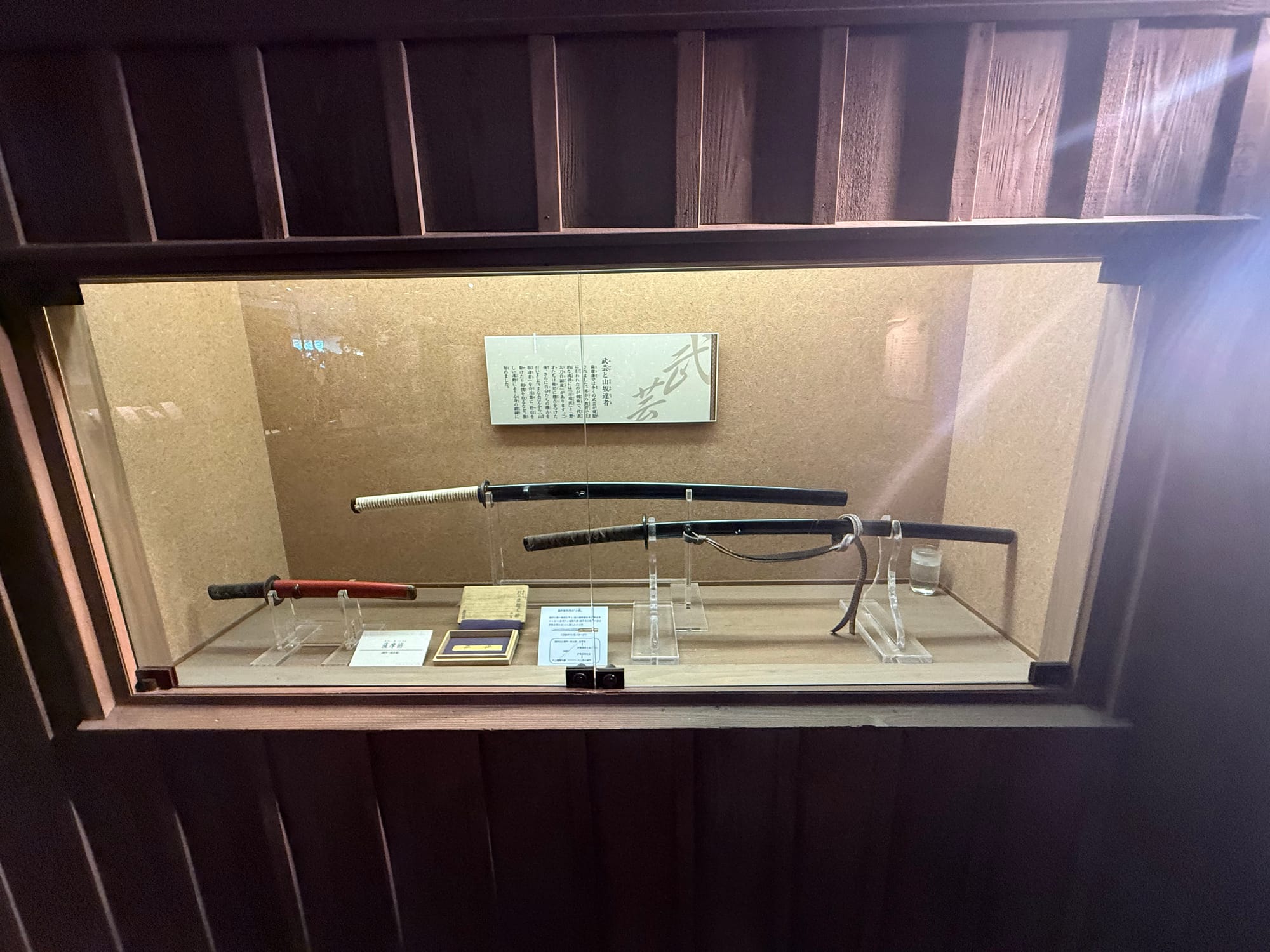 Display case at the Museum of the Meiji Restoration showcasing Japanese swords and their scabbards, including a katana and wakizashi, mounted on stands with explanatory labels.