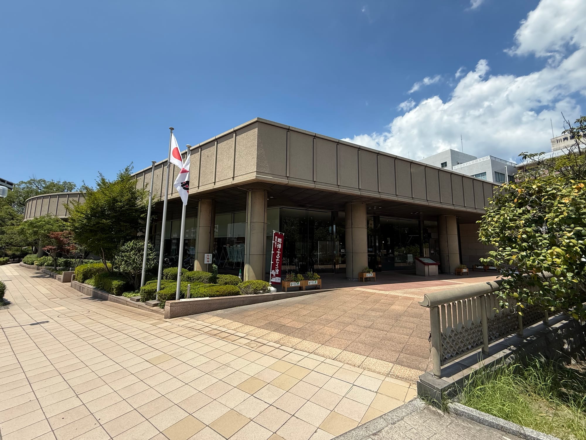 Museum of the Meiji Restoration in Kagoshima, a modern museum building with a colonnaded entrance, Japanese flags and landscaped grounds beneath a bright blue sky.