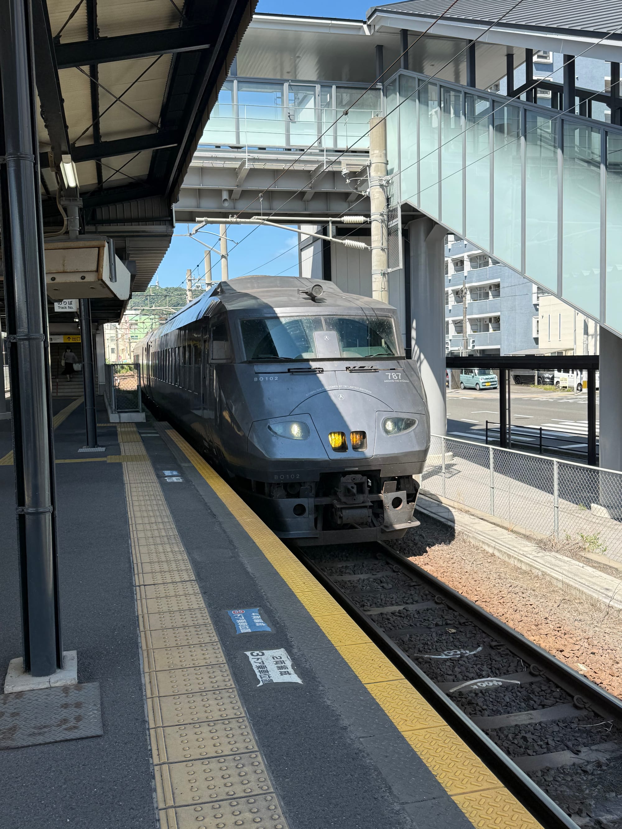 JR Kyushu 787 Series limited express train standing at a platform in Kagoshima, with its distinctive dark-grey livery and streamlined front beneath a station footbridge.