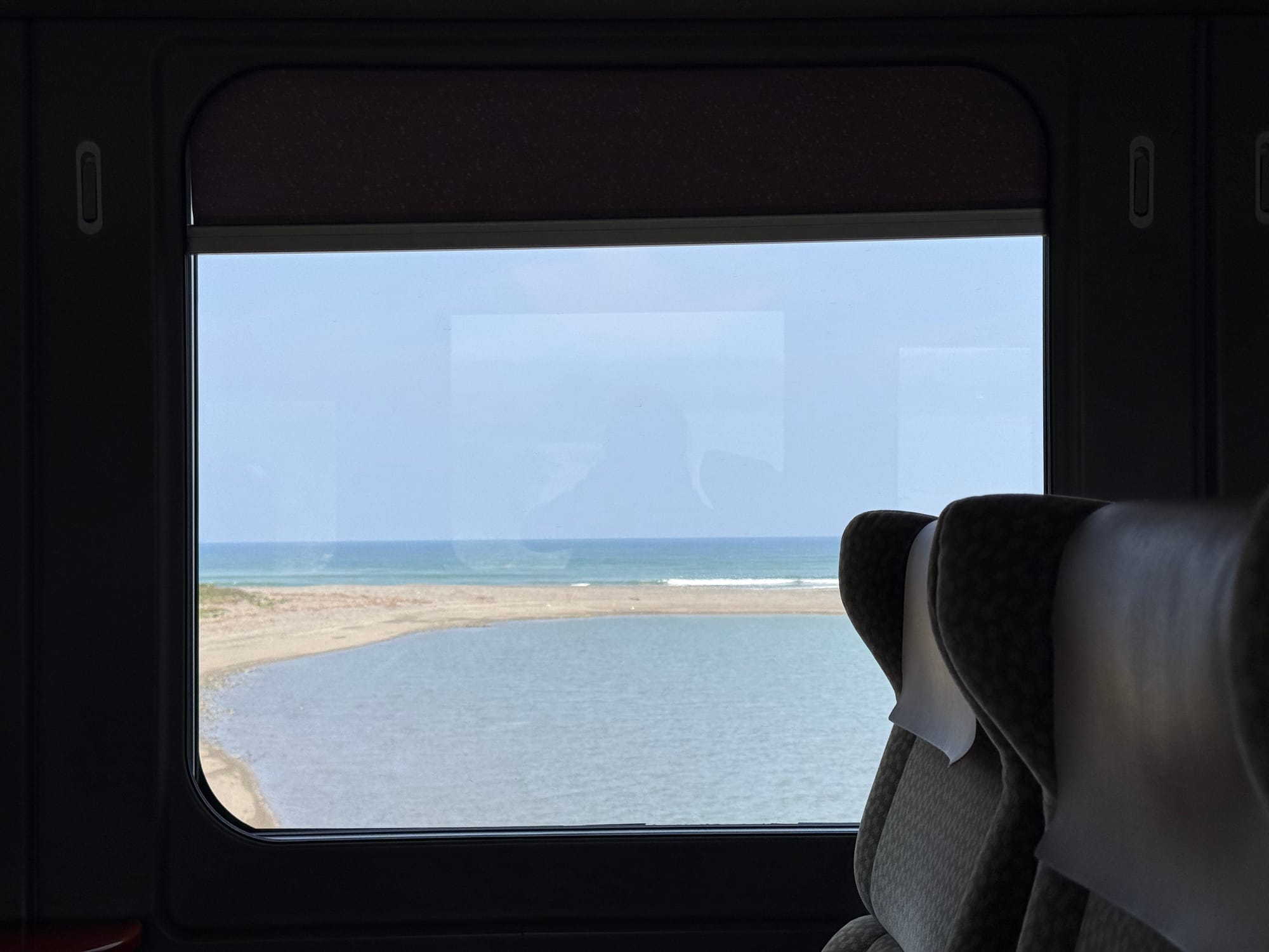 View from a JR Kyushu 787 Series train window, looking across a coastal lagoon, sandy shoreline and the ocean beyond, with Green Car seats visible in the foreground.