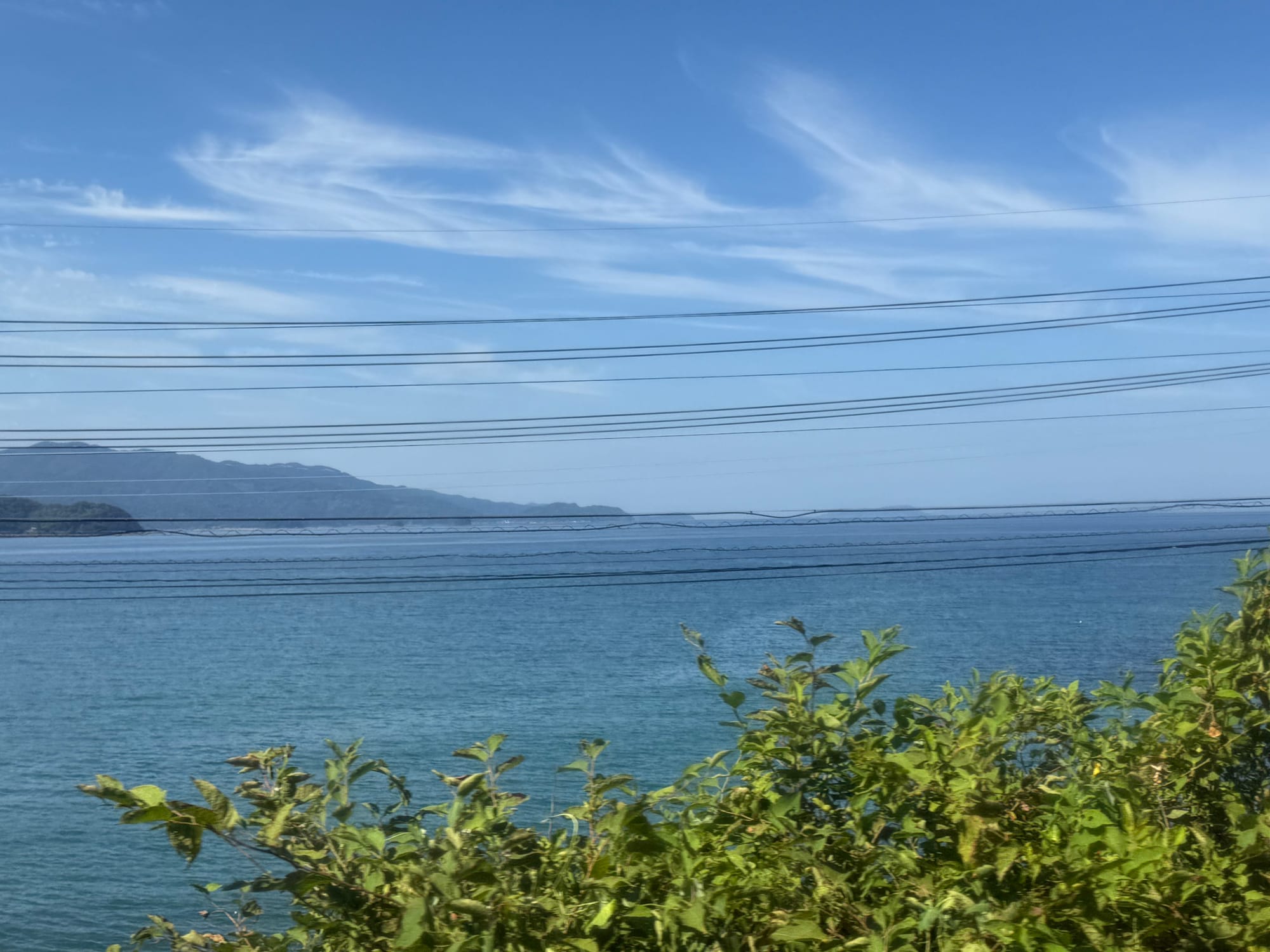 View from a JR Kyushu 787 Series train window showing turquoise coastal waters, a distant headland and wispy clouds above the sea along Kyushu's eastern shoreline.