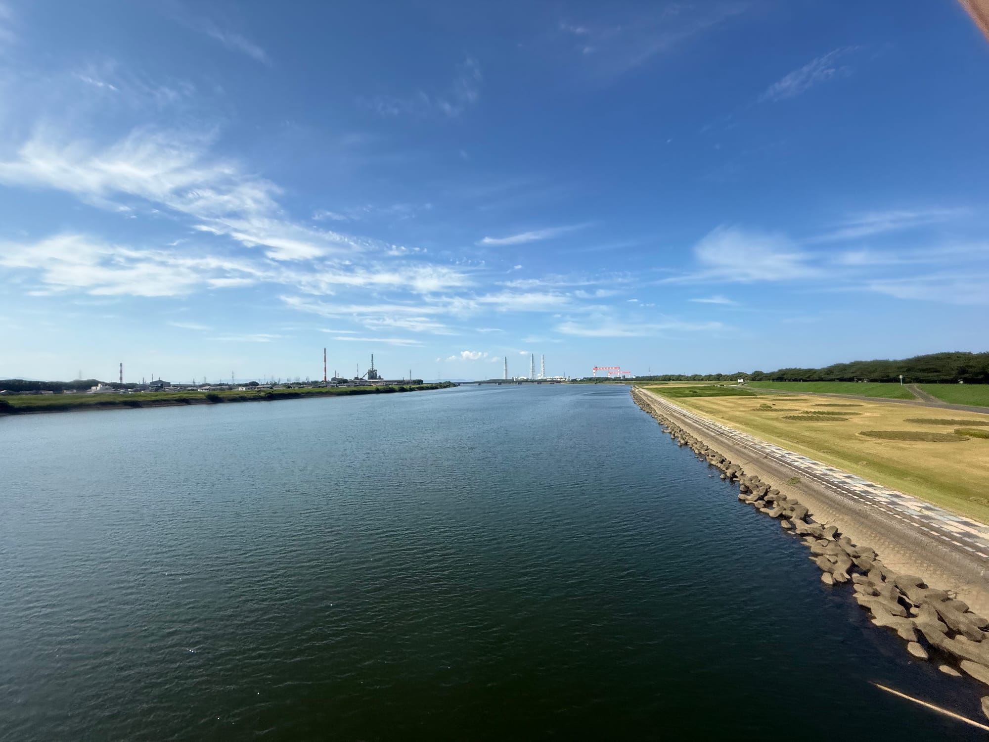 View from a train crossing a wide river in Kyushu, with calm water, embankments, distant industrial facilities and high clouds stretching across a bright blue sky.