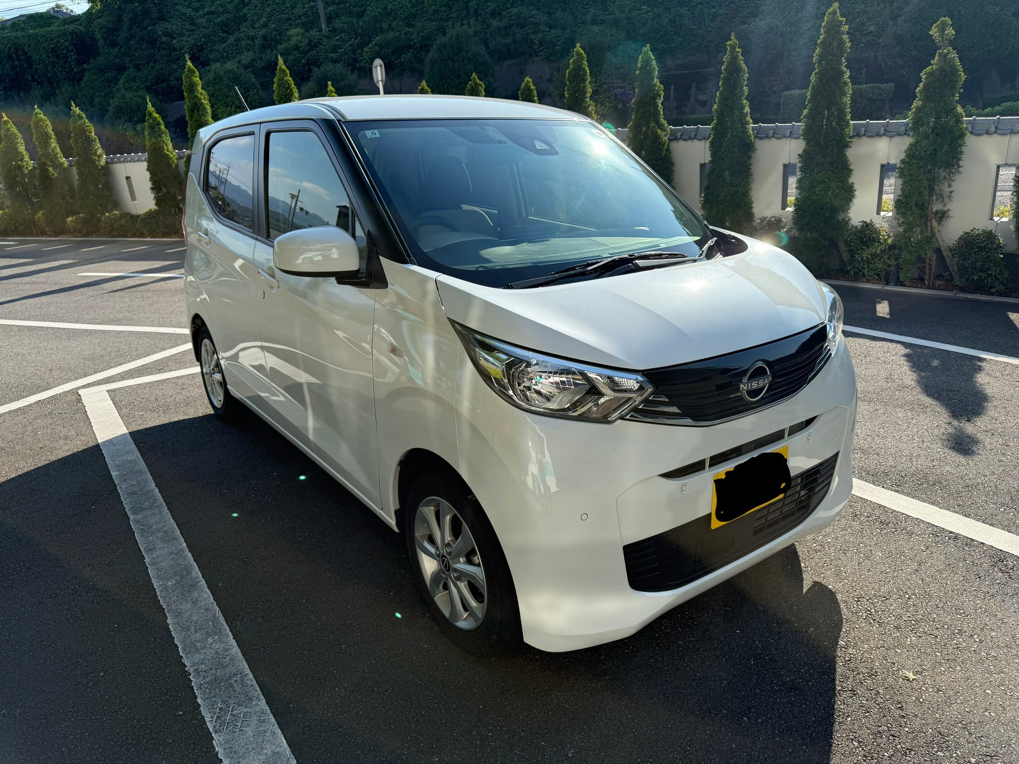 White Nissan Dayz hire car parked in a car park near Beppu, with afternoon sunlight reflecting from the bodywork and forested hills rising in the background.