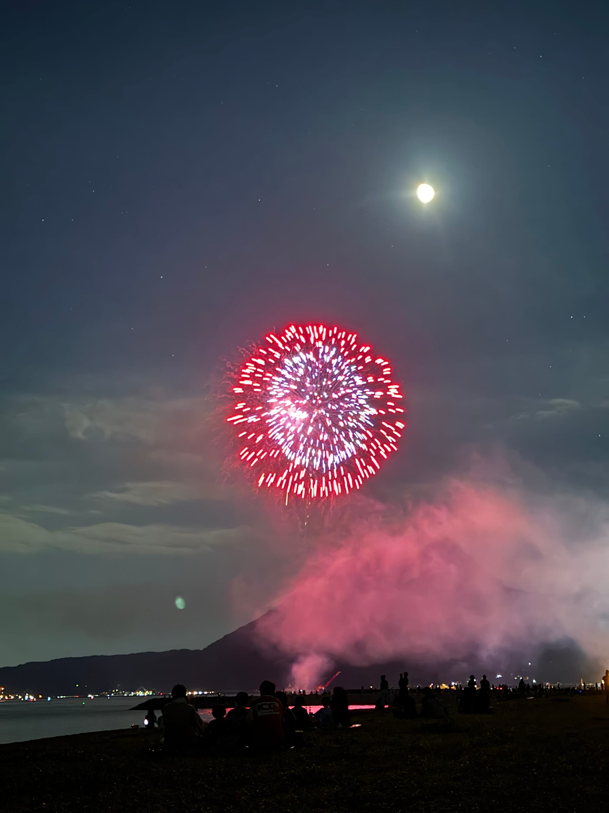 Fireworks explode above the shoreline at night, with spectators watching beneath a bright moon as smoke drifts across the silhouette of a coastal mountain.