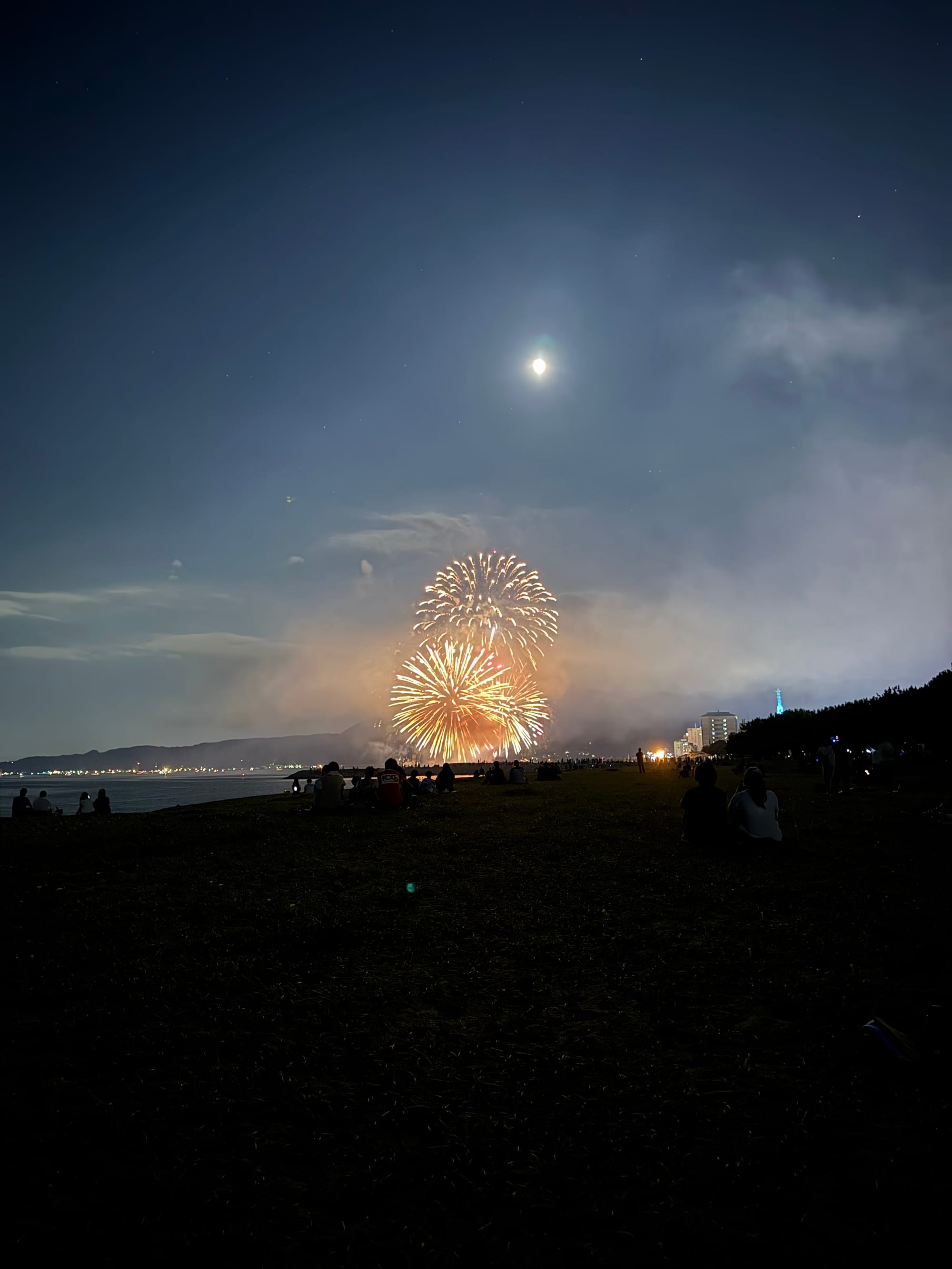 Fireworks burst above the shoreline at night, illuminating drifting smoke as spectators watch from the beach beneath a bright moon and a star-filled sky.