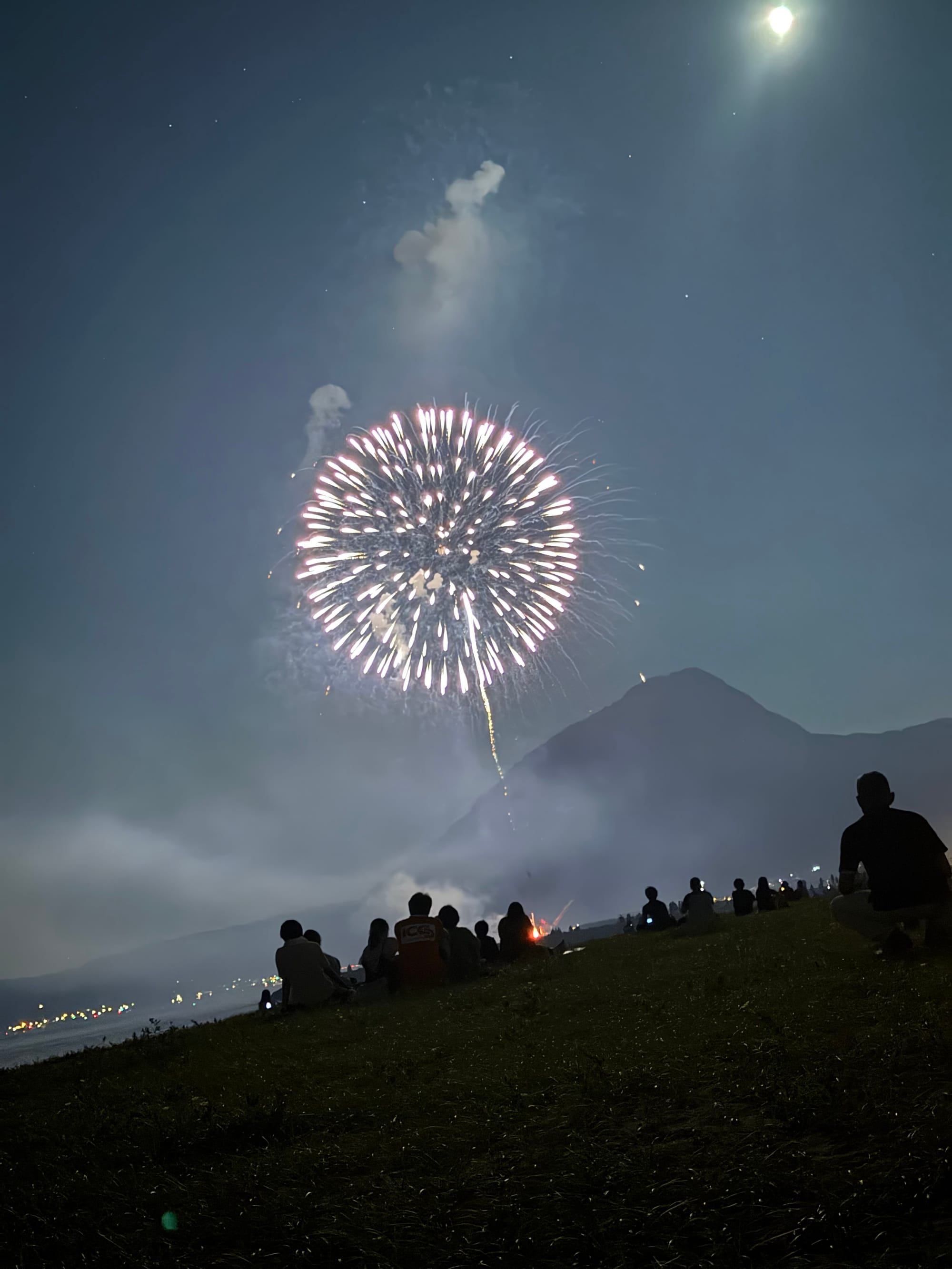 White firework bursts above a coastal hillside at night, with spectators seated on the grass below and a bright moon shining through drifting smoke.