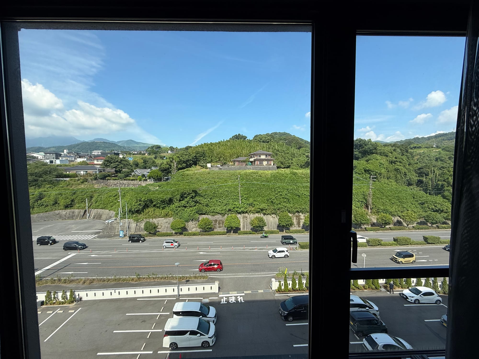 View from a hotel room window in Beppu, overlooking a main road, parked cars, green hillsides and scattered houses beneath a bright blue summer sky.
