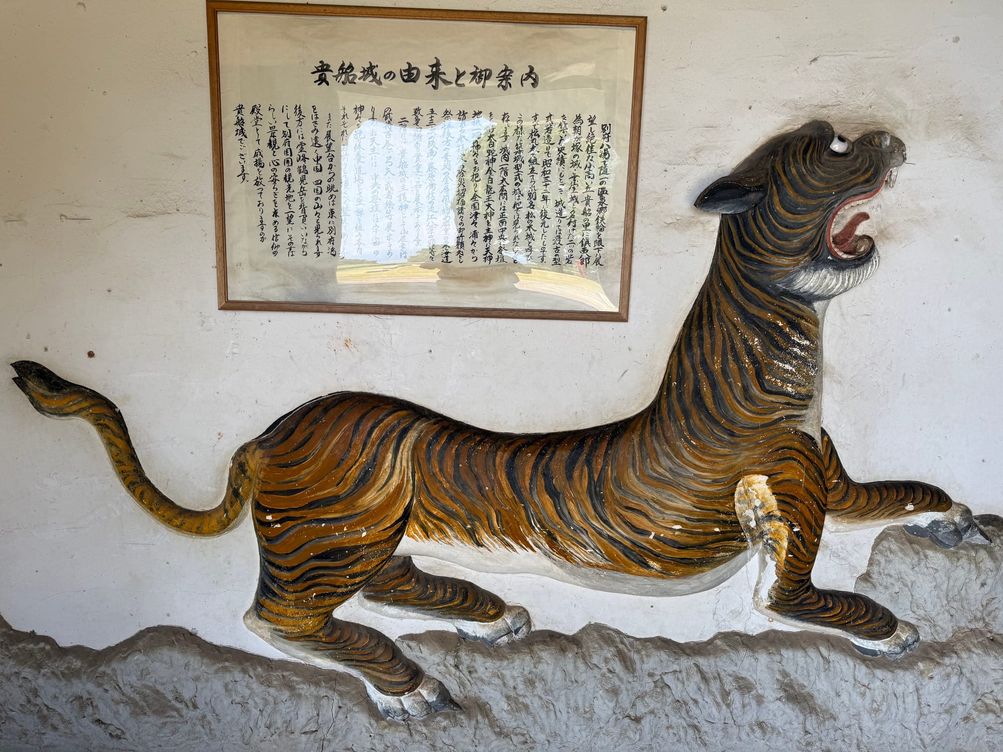Relief sculpture of a tiger mounted on a wall at Kifune Castle, shown in mid-stride beneath a framed panel of Japanese text explaining part of the castle's history.