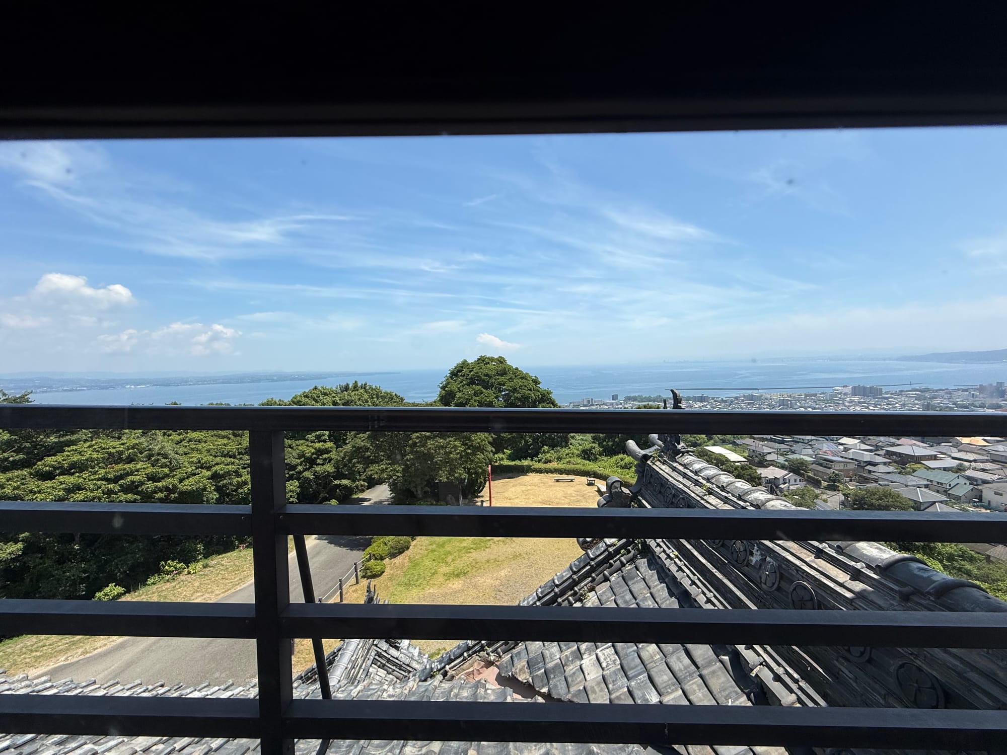 View from the upper level of Kifune Castle across Beppu and Beppu Bay, with traditional tiled rooftops, trees, the coastline and calm blue water beneath a clear sky.