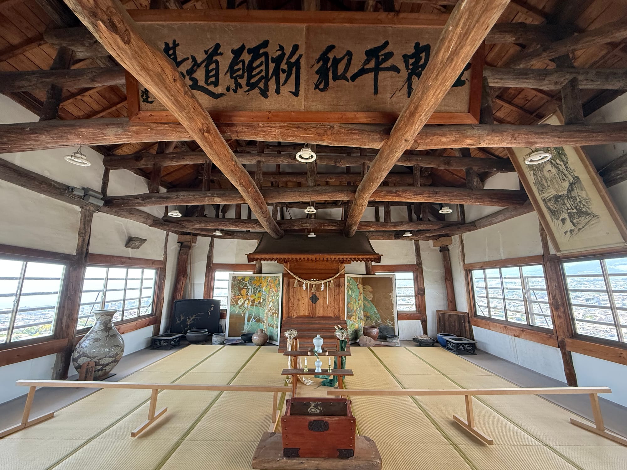 Top floor of Kifune Castle featuring tatami flooring, exposed timber beams, decorative screens, ceremonial objects and panoramic windows overlooking Beppu.