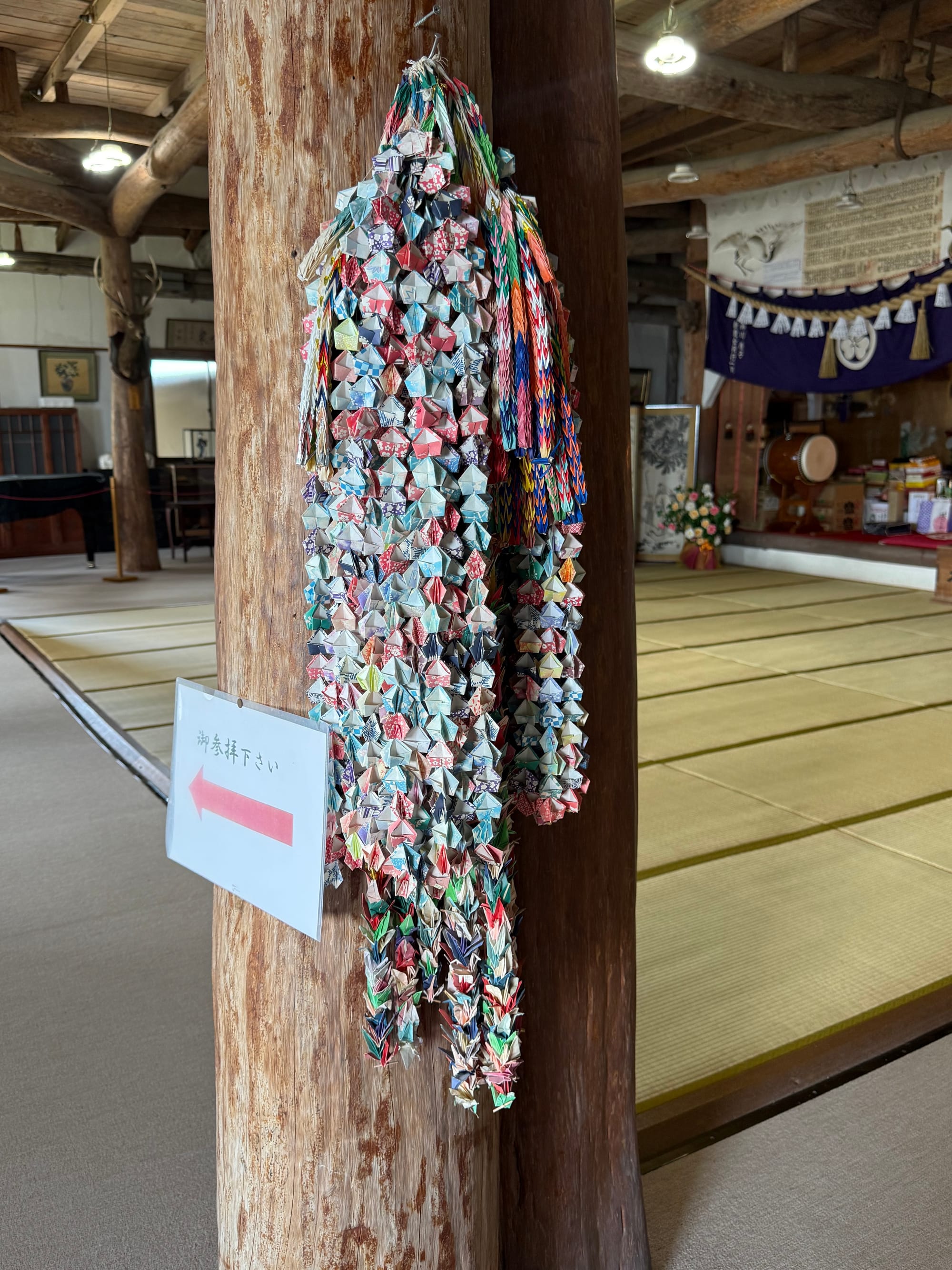 Colourful strings of folded origami cranes hang from a wooden pillar inside Kifune Castle, displayed among tatami-floored rooms, timber beams and traditional décor.