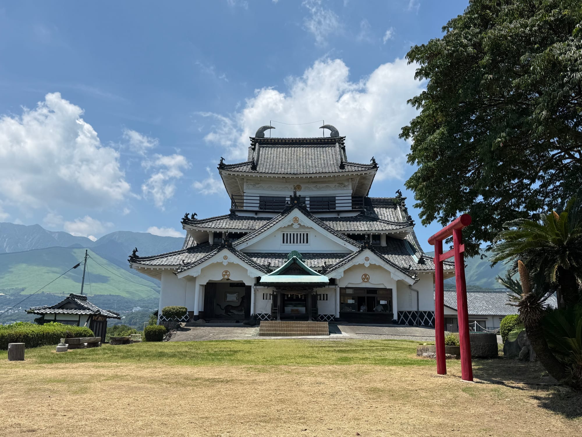 Kifune Castle near Beppu, featuring white plaster walls, black tiled roofs and a red torii gate, with the mountains of Ōita Prefecture rising behind beneath a blue sky.