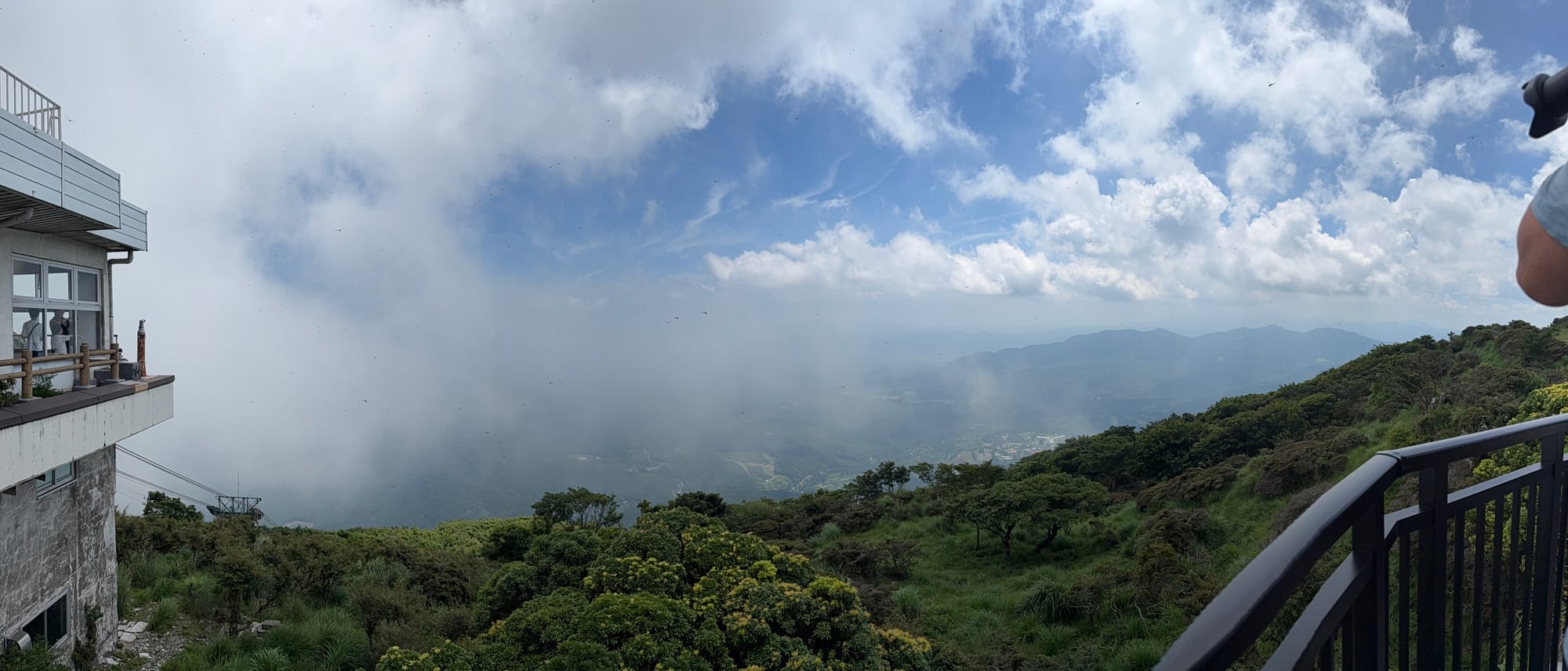 View from the upper station of the Beppu Ropeway on Mount Tsurumi, with clouds drifting across forested slopes and distant mountain ridges beneath a bright summer sky.