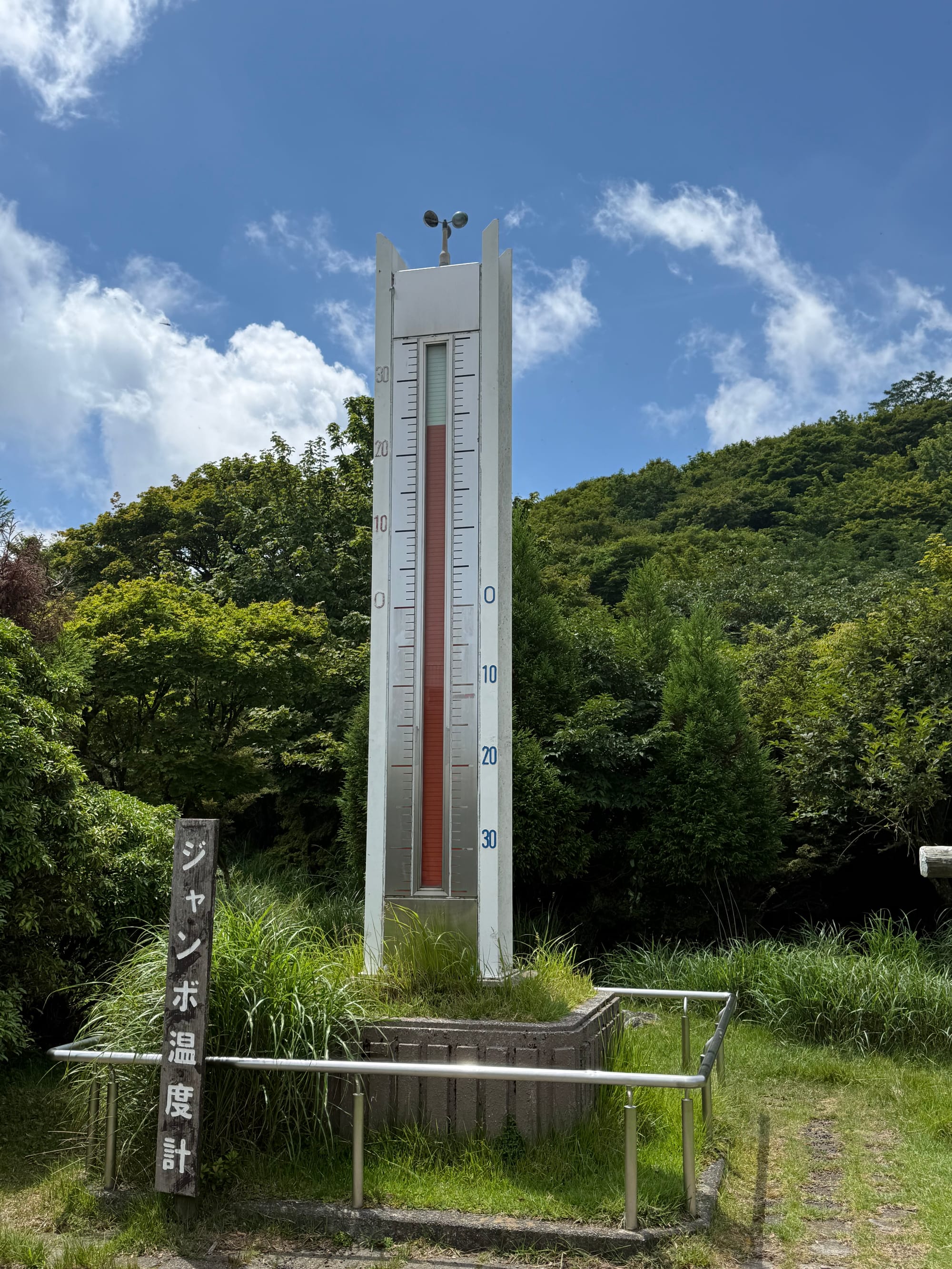 Large outdoor thermometer at the summit of Mount Tsurumi near Beppu, showing the current temperature against a backdrop of forested slopes and blue summer skies.