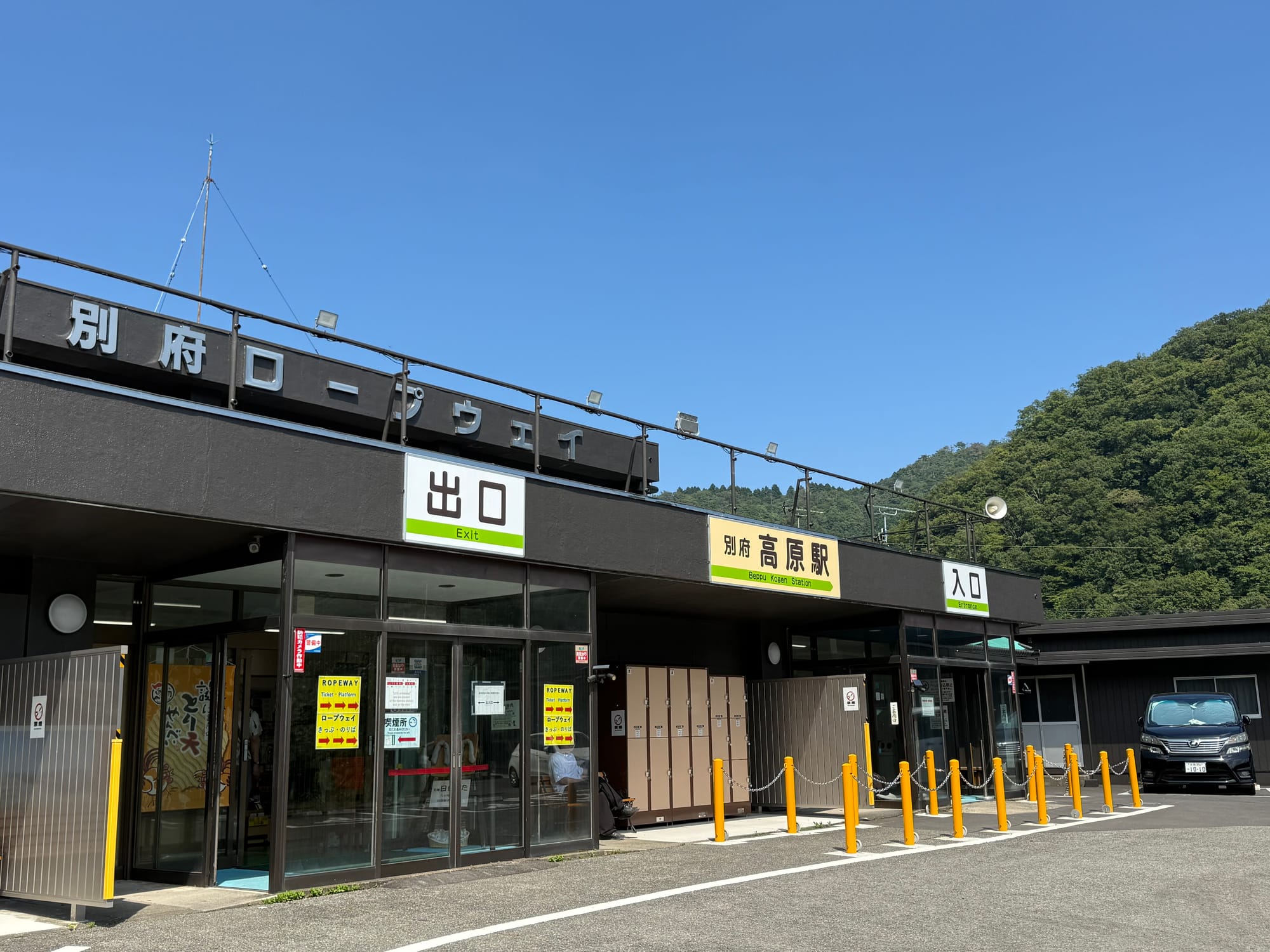 Beppu Kōgen Station building, featuring bilingual station signage, glass entrance doors and forested hills behind, beneath a clear blue summer sky.