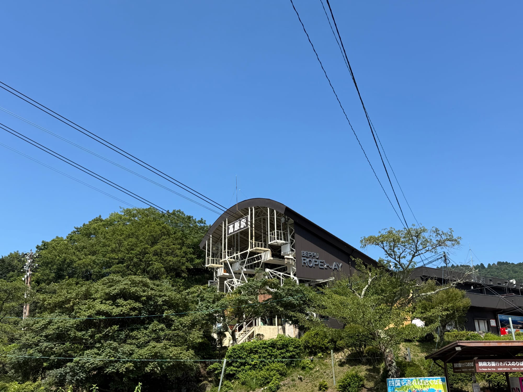 Lower station of the Beppu Ropeway, nestled among forested hills, with the ropeway terminal building and support structures visible beneath a clear blue summer sky.