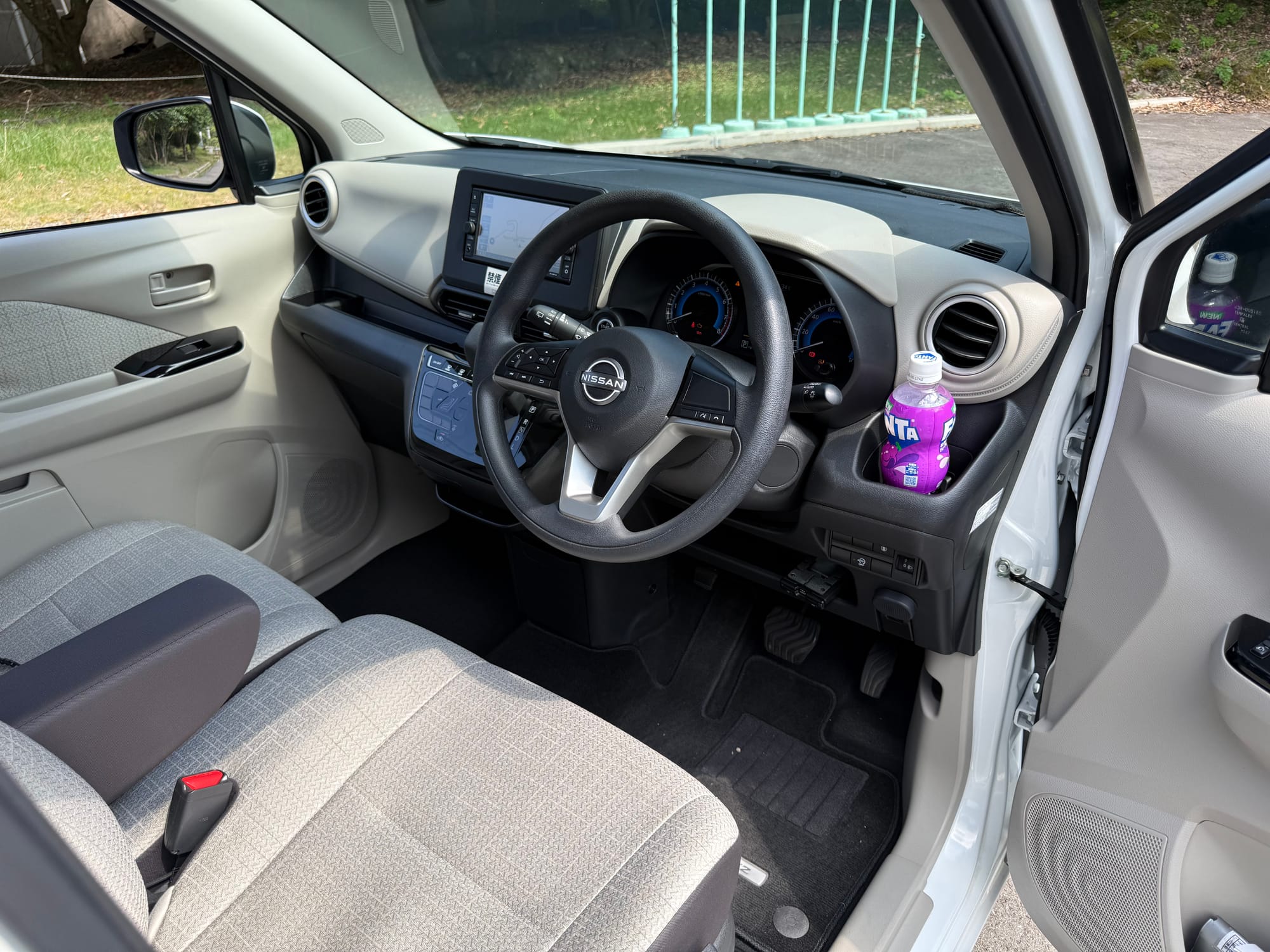 Interior of a Nissan Dayz hire car in Beppu, showing the right-hand-drive layout, dashboard display, fabric seats and a bottle of grape Fanta in the door pocket.