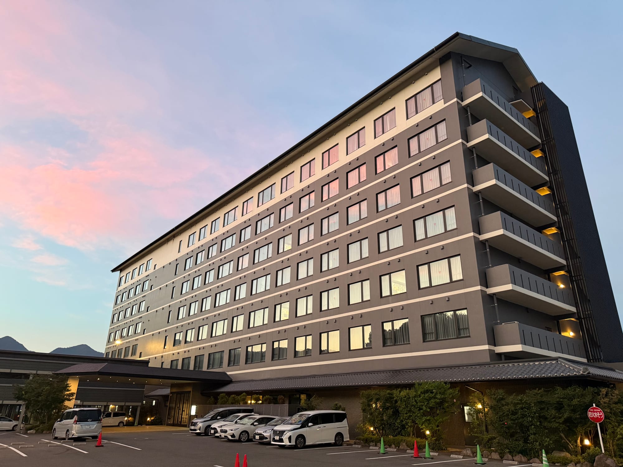 Large modern hotel in Beppu at sunset, with rows of illuminated windows, parked cars and soft pink clouds reflected in the glass façade beneath the evening sky.