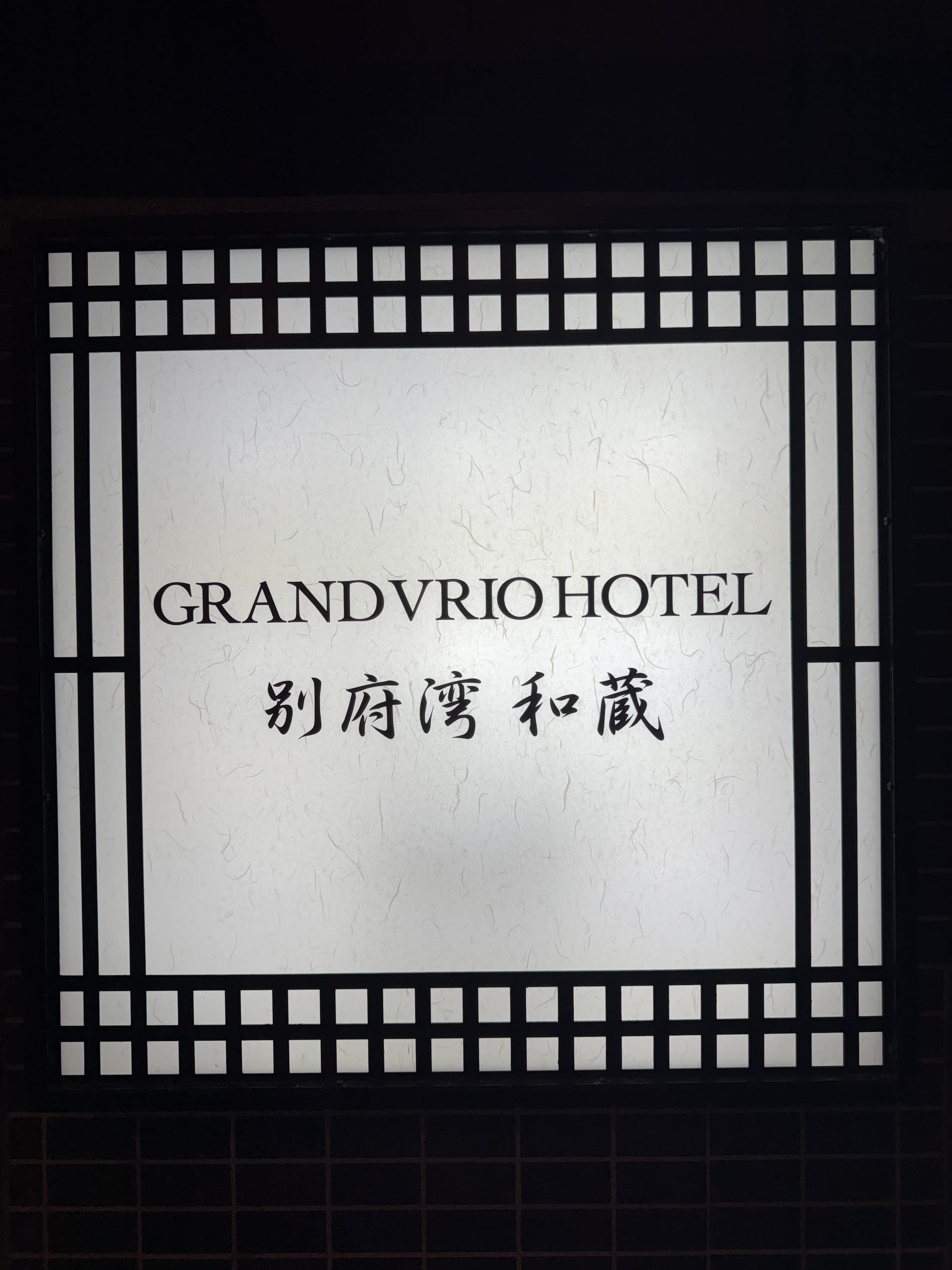 Illuminated sign for Grandvrio Hotel Beppu Bay Wakura, displaying the hotel name in English and Japanese against a traditional-style white-and-black backdrop at night.