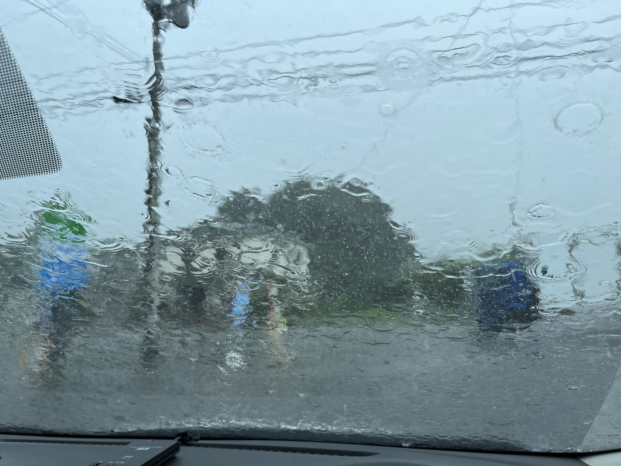 View through the windscreen of a Nissan Note during heavy rain, parked at a Japanese convenience store, with water streaming across the glass and blurring the scene outside.