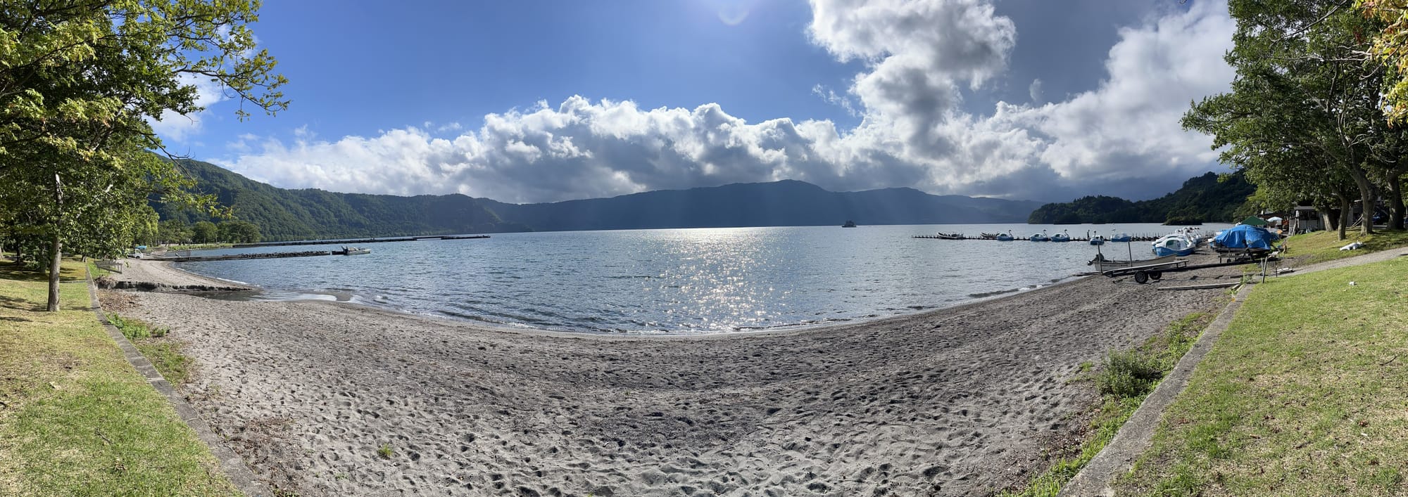 Panoramic view of Lake Towada with a sandy lakeshore, wooded hills and boats on the water, illuminated by sunlight breaking through dramatic clouds.