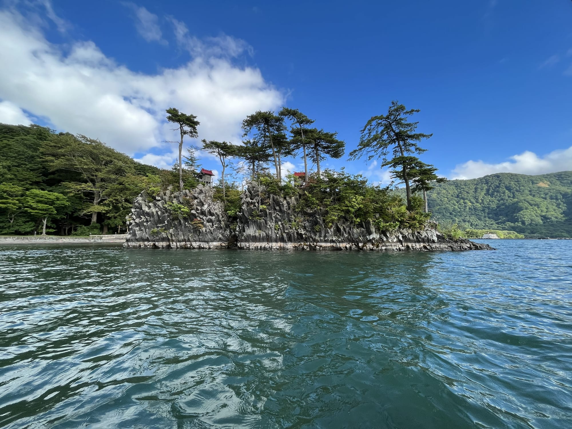 Rocky island in Lake Towada topped with windswept pine trees and small shrine structures, rising above clear water beneath a bright blue sky.