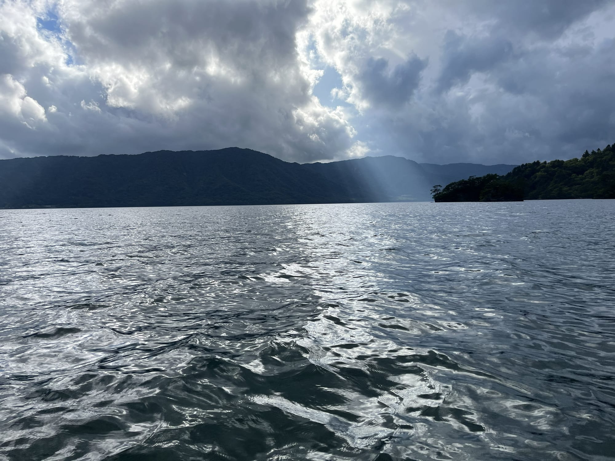Sunbeams break through dramatic clouds above Lake Towada, illuminating the water and surrounding forested mountains after a passing rain shower.