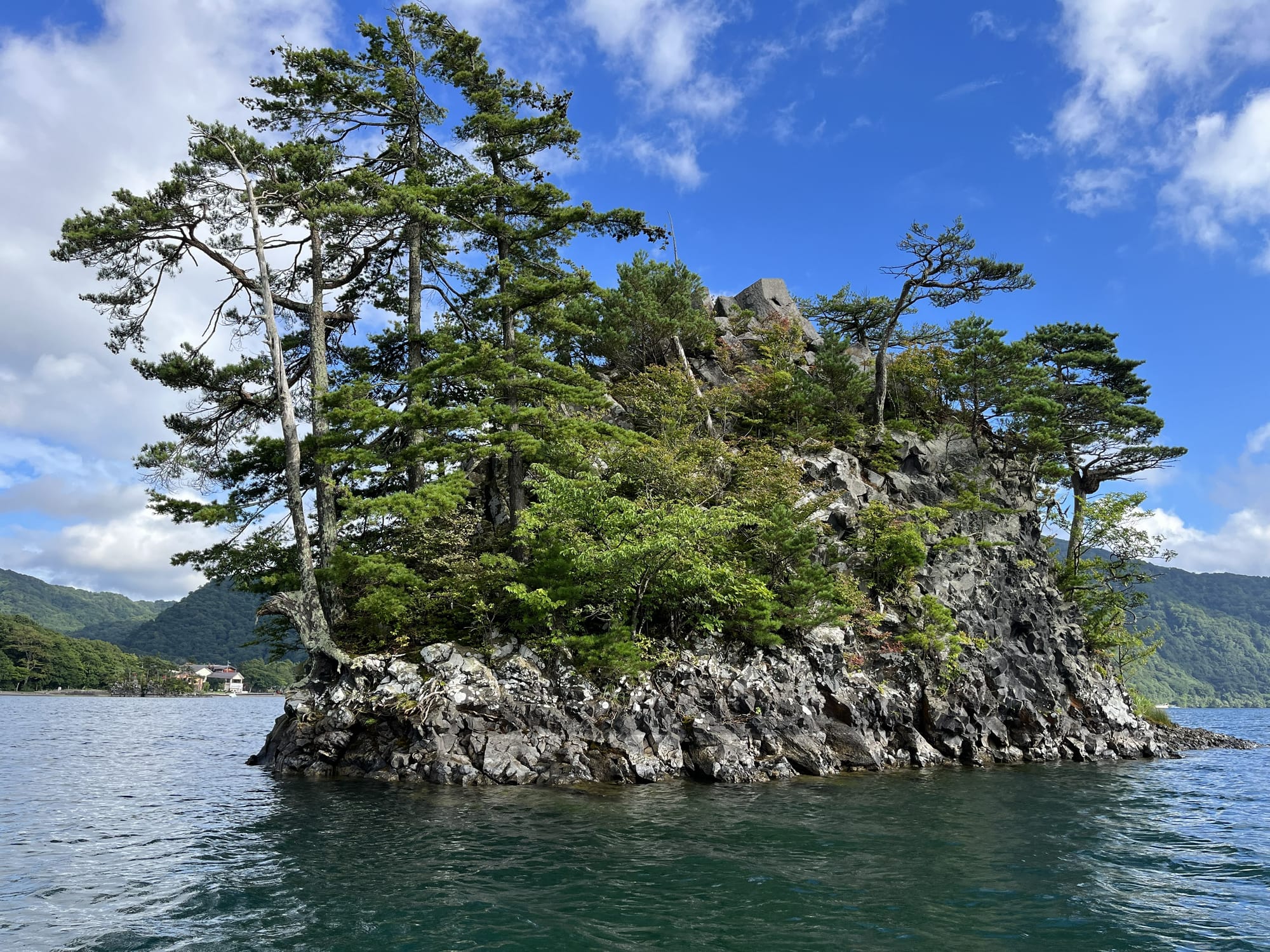 Rocky, tree-covered island in Lake Towada, with windswept pines growing from steep volcanic cliffs above clear blue water beneath a bright sky.