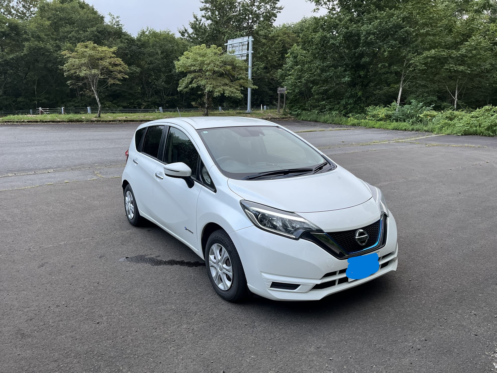 White Nissan Note hire car parked in a roadside lay-by surrounded by forest on the route to Lake Towada, photographed on an overcast day in northern Japan.