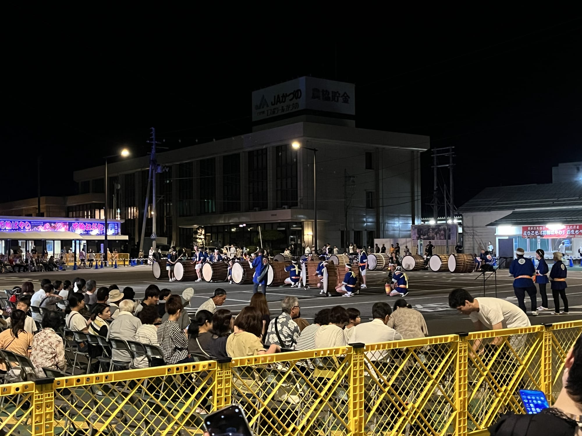 Traditional taiko drummers perform in an outdoor festival in Kazuno at night, with spectators seated behind barriers as large drums line the performance area.