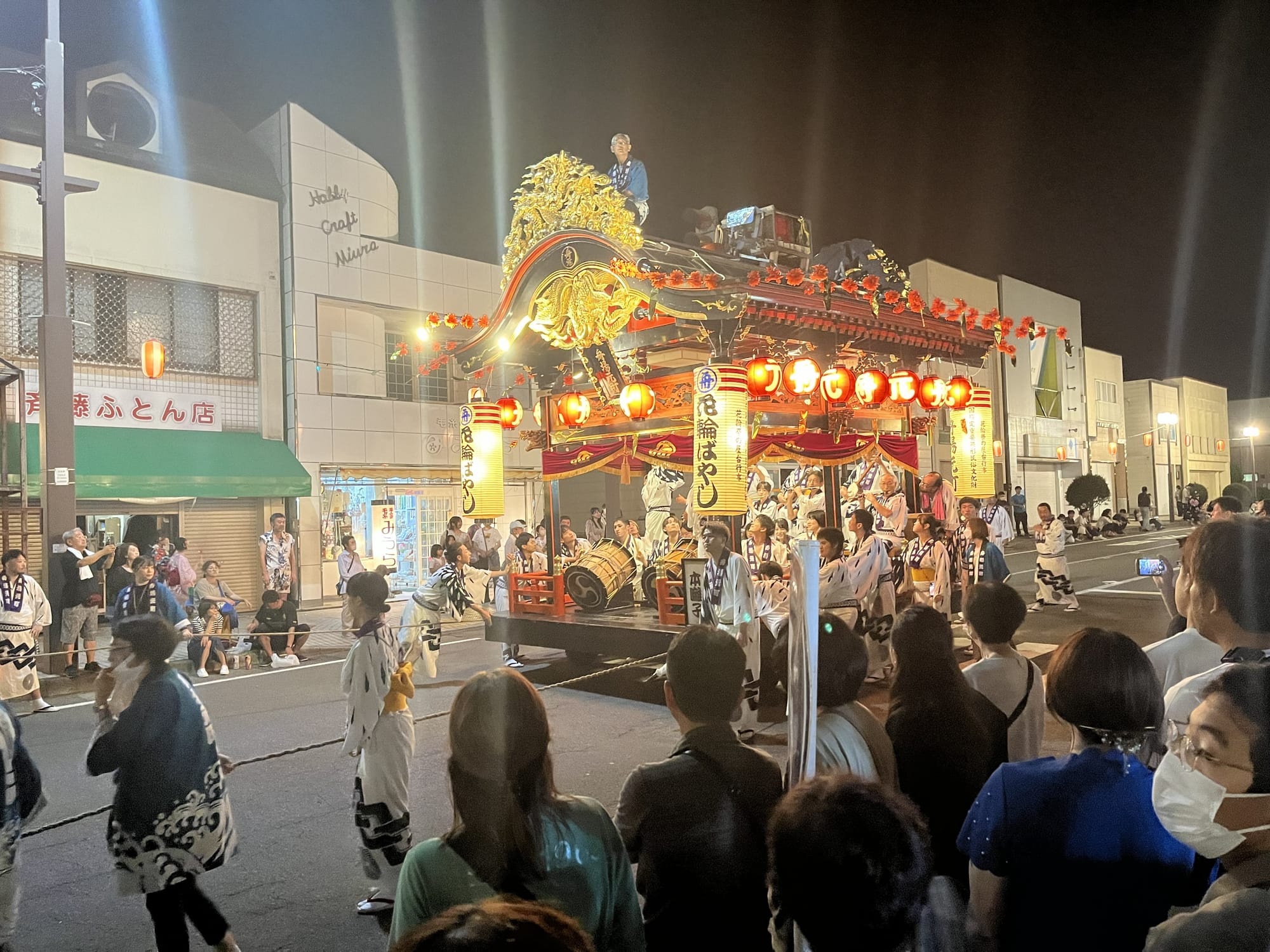 Illuminated festival float decorated with lanterns and ornate carvings moves through the streets of Kazuno at night, surrounded by spectators and performers in traditional dress.