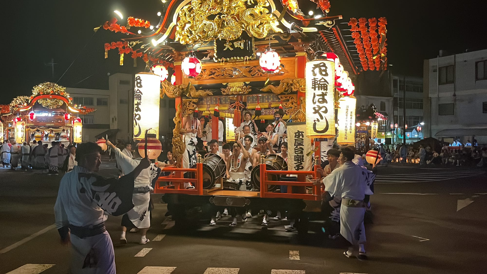 Illuminated festival float adorned with lanterns and ornate gold carvings moves through the streets of Kazuno at night, accompanied by drummers and participants in traditional dress.
